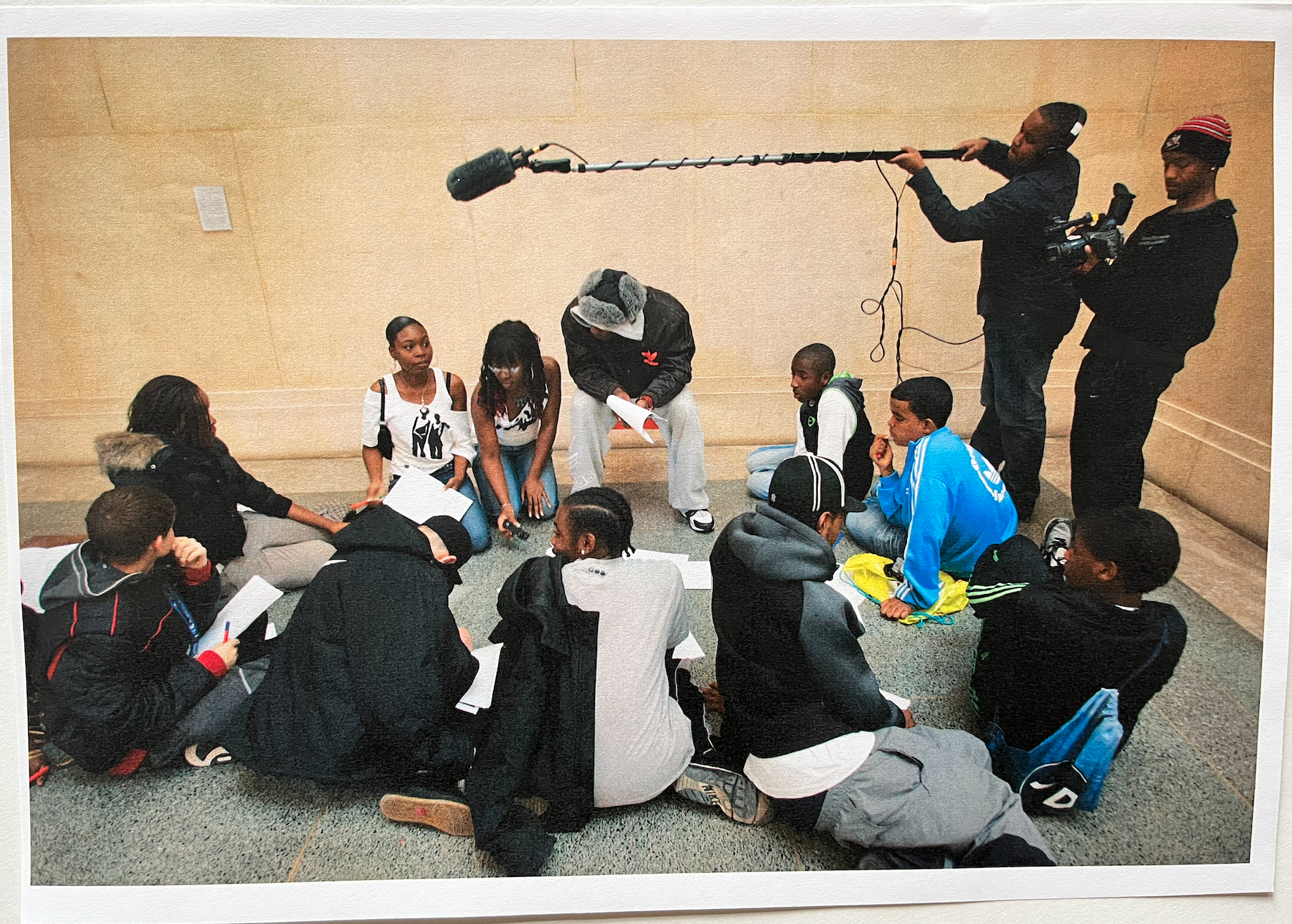 Group of 10 young people sitting on the floor of a gallery looking focused on conversation with pieces of paper and pens. 2 young people with filming and recording devices capture the group.