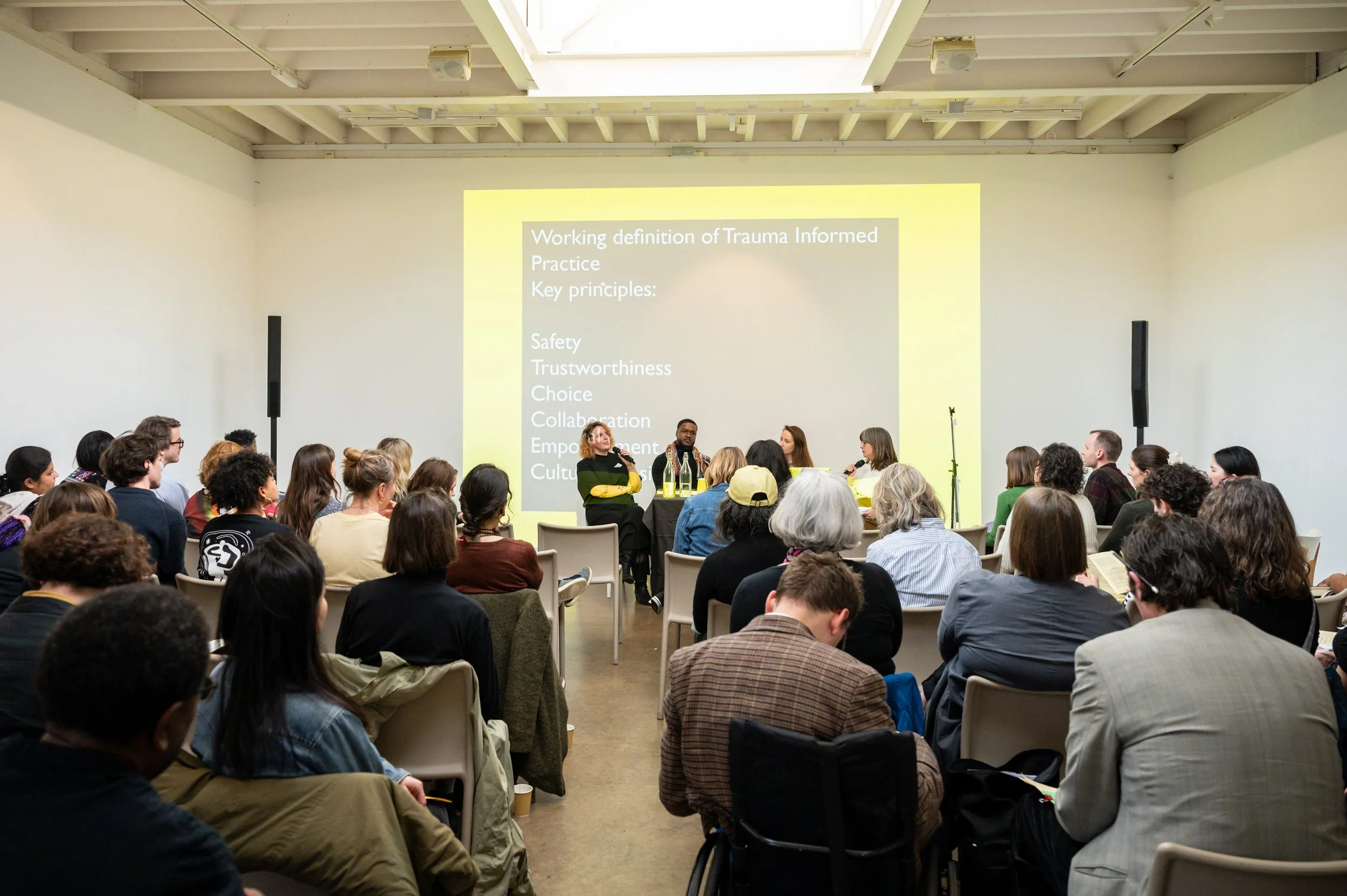 Large audience of 100 people listening to 4 adults in conversation. Slide projected in background says 'Working definition of trauma informed practice' to