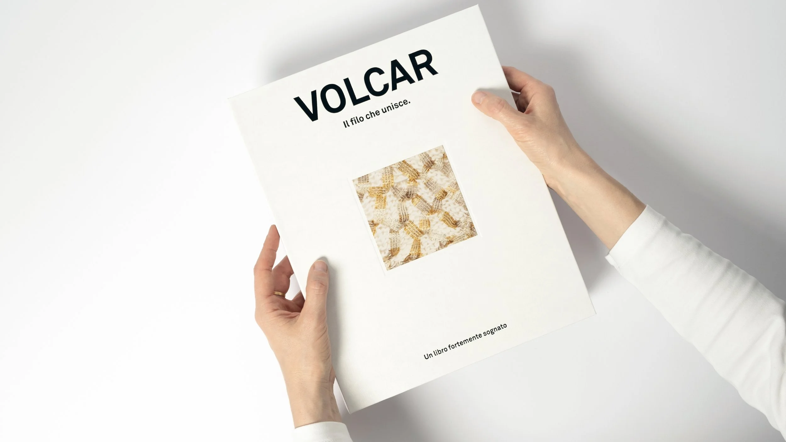 Una persona tiene tra le mani un libro intitolato 'VOLCAR. Il filo che unisce', che mostra sulla copertina un'immagine di un tessuto intrecciato oro e bianco, con il sottotitolo 'Un libro fortemente sognato'.