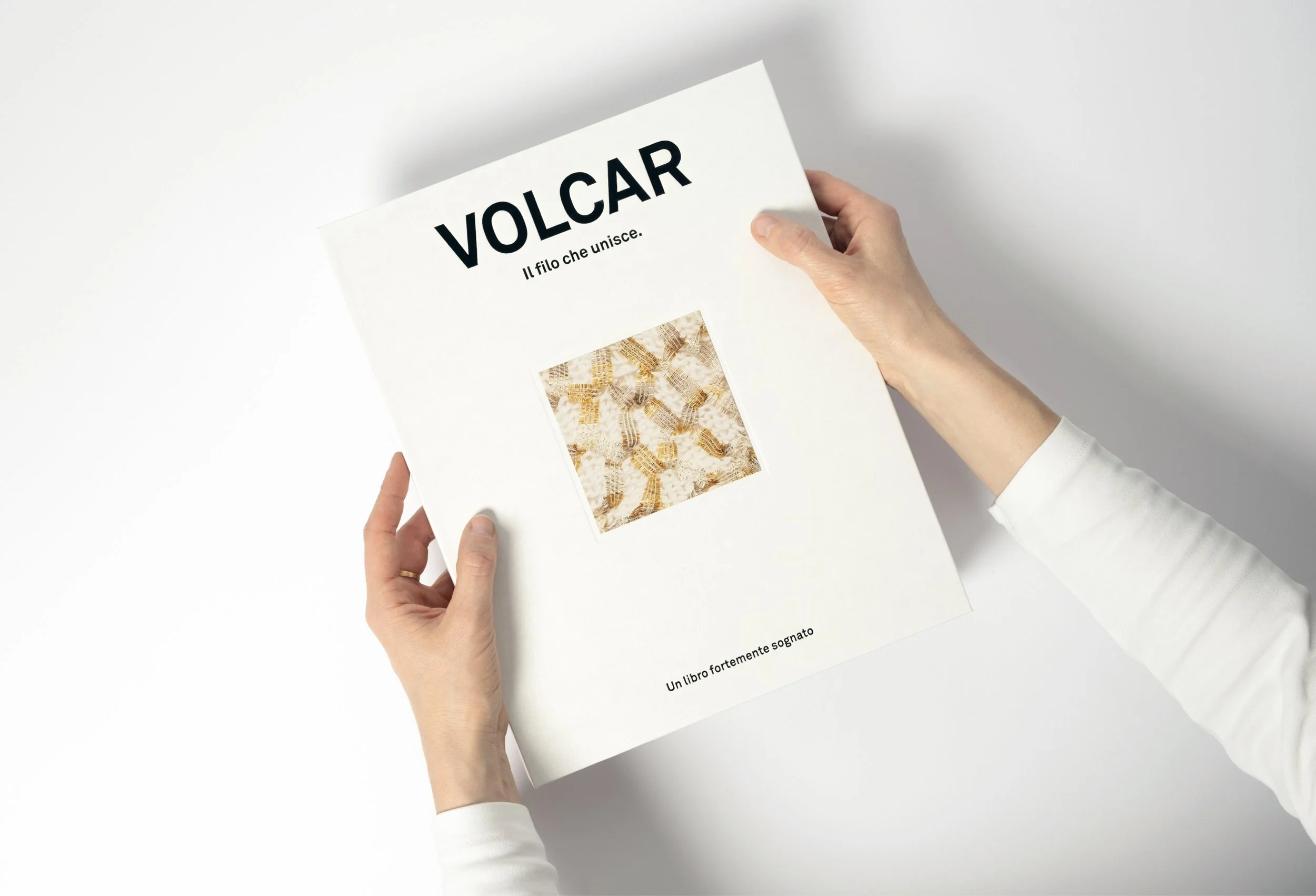 Una persona tiene tra le mani un libro intitolato 'VOLCAR. Il filo che unisce', che mostra sulla copertina un'immagine di un tessuto intrecciato oro e bianco, con il sottotitolo 'Un libro fortemente sognato'.