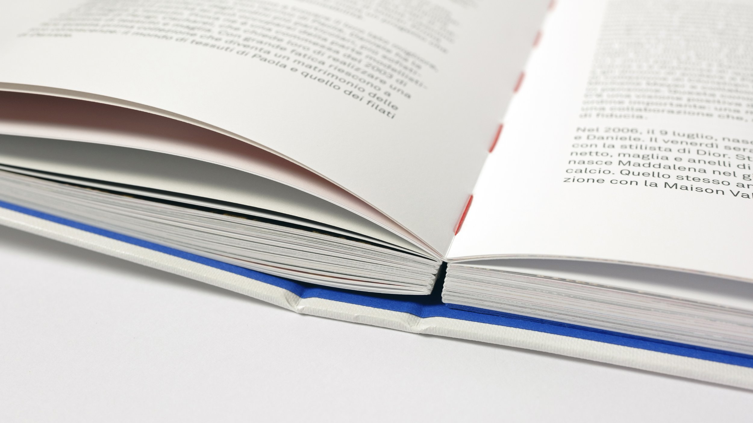 Particolare di un libro aperto con pagine bianche e testo scritto. Si vede la cucitura a filo in cotone color rosso.