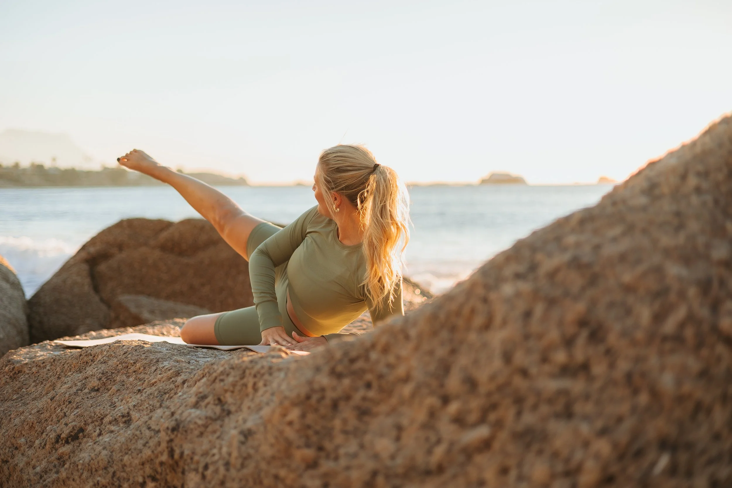 A woman practicing yoga on rocks by the beach during sunset.
