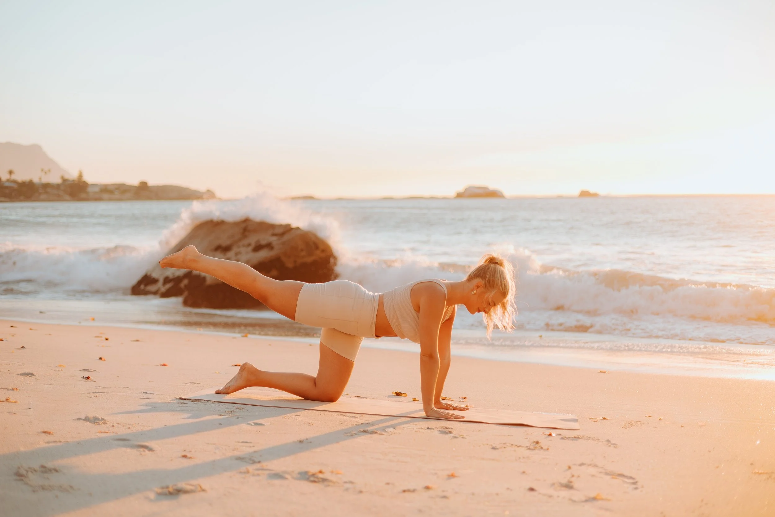 Woman practicing yoga on the beach at sunset, in a modified push-up position with one leg raised, wearing workout clothes.