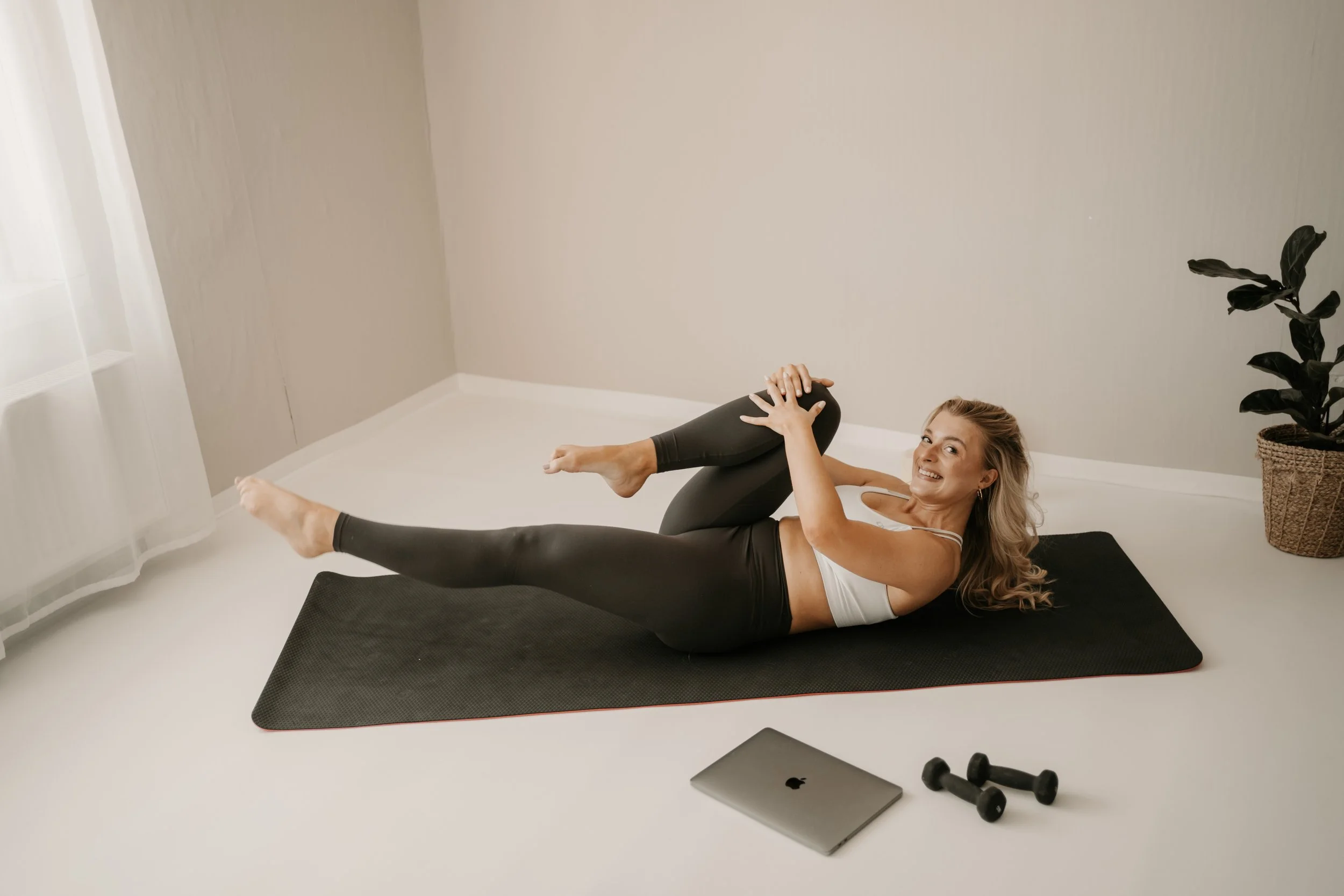Woman lying on a yoga mat in a bright room, smiling and holding her leg while doing a stretching exercise. There are two dumbbells, a laptop, and a potted plant in the background.