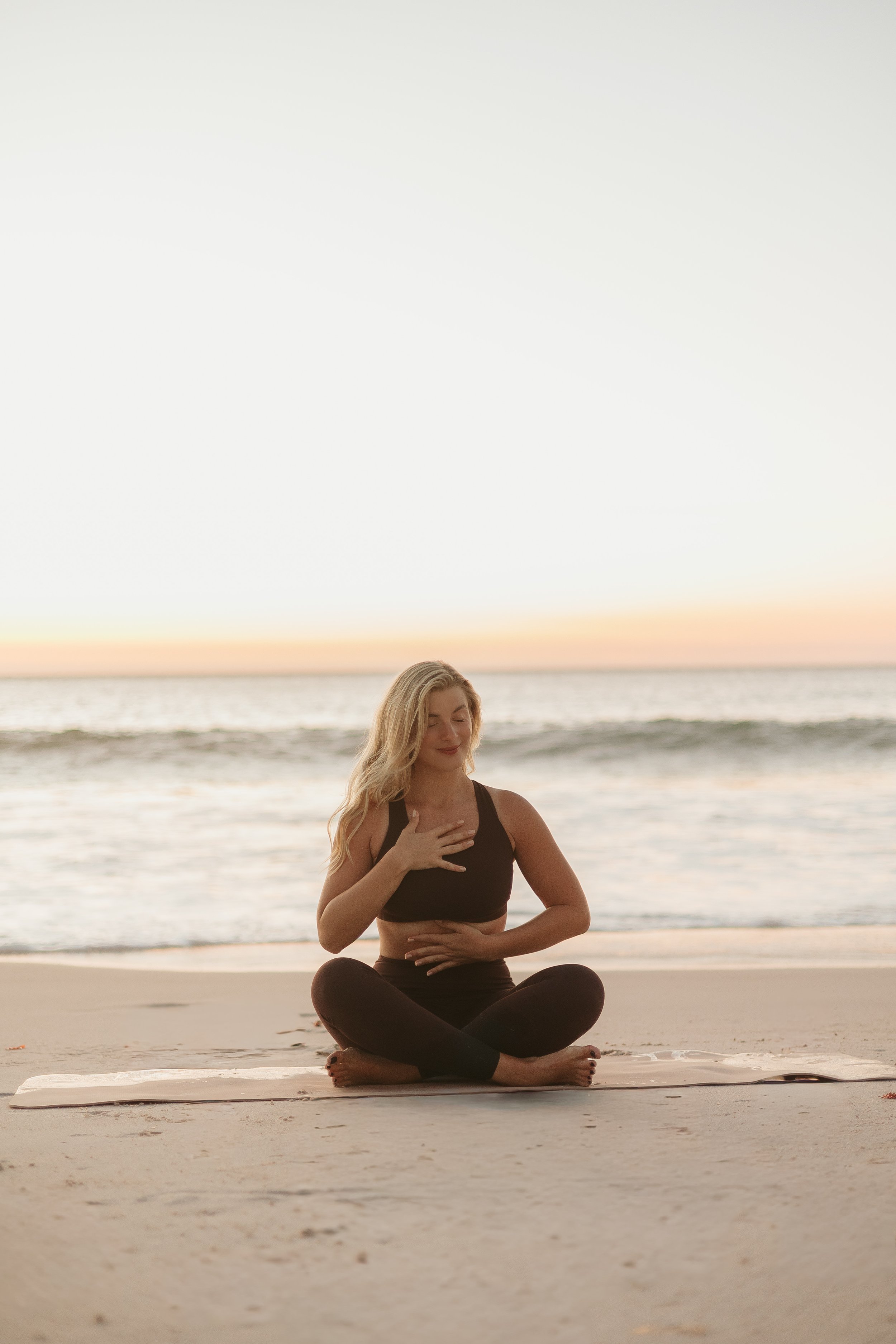 A woman practicing yoga on the beach at sunset, sitting cross-legged with one hand on her chest and the other on her stomach.