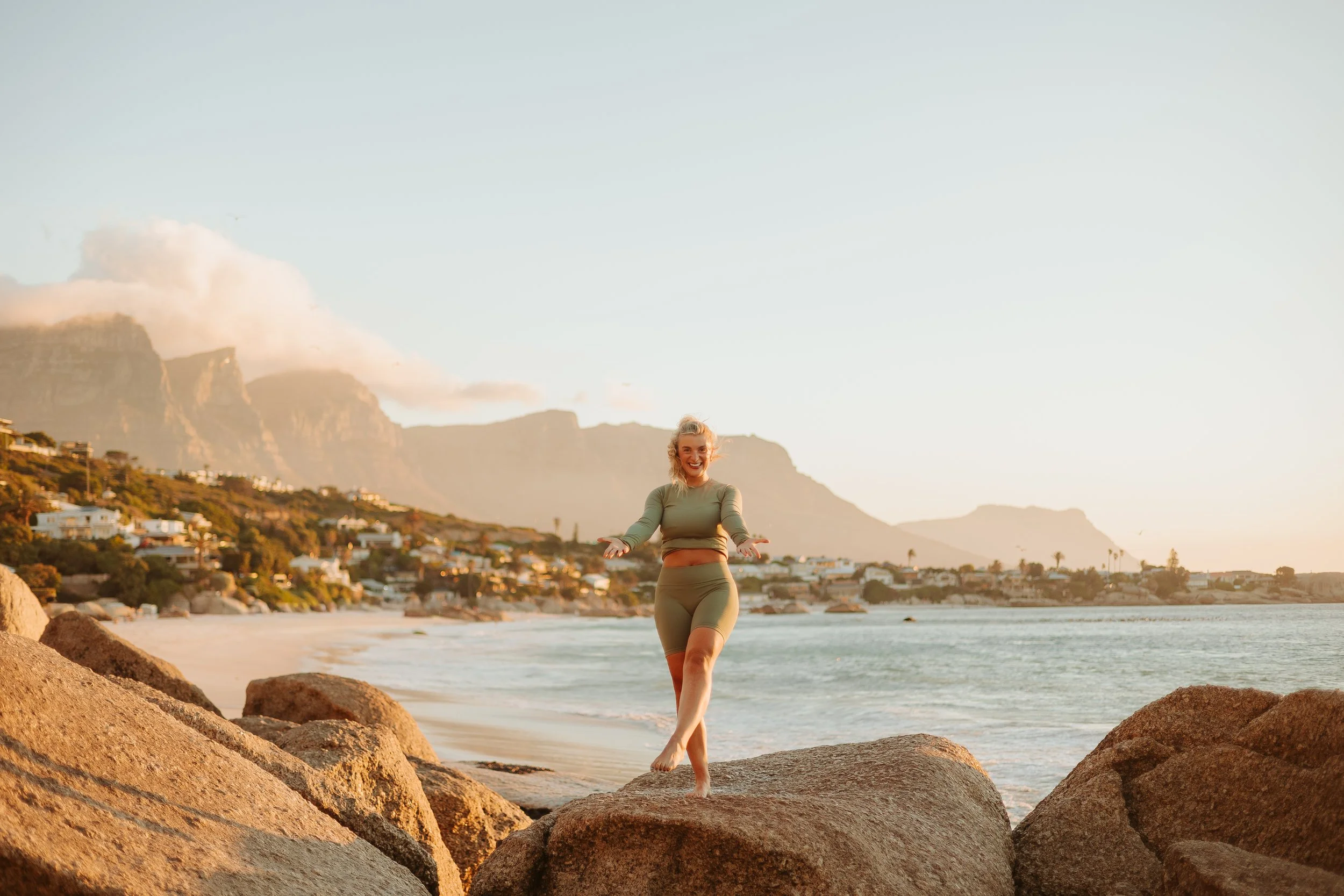 A woman standing on rocks near the beach with a shoreline, houses, and mountains in the background during sunset.