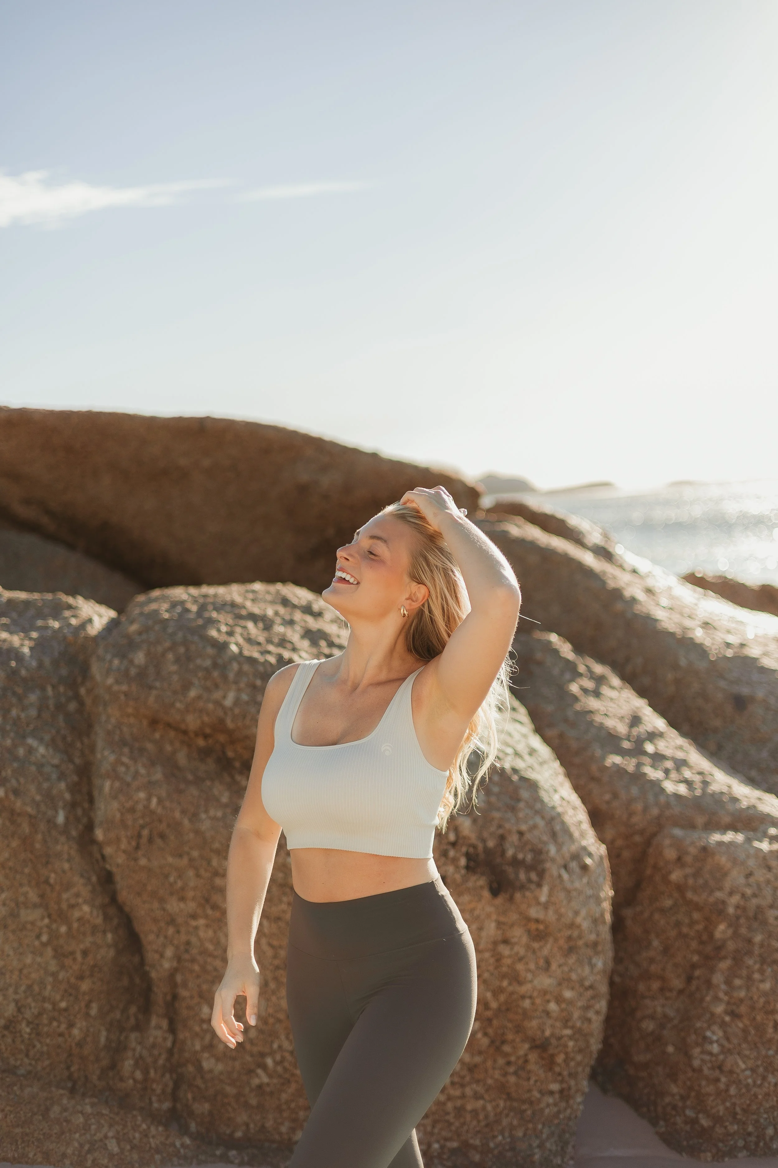 A woman smiling and enjoying sunshine on a beach with large rocks in the background.