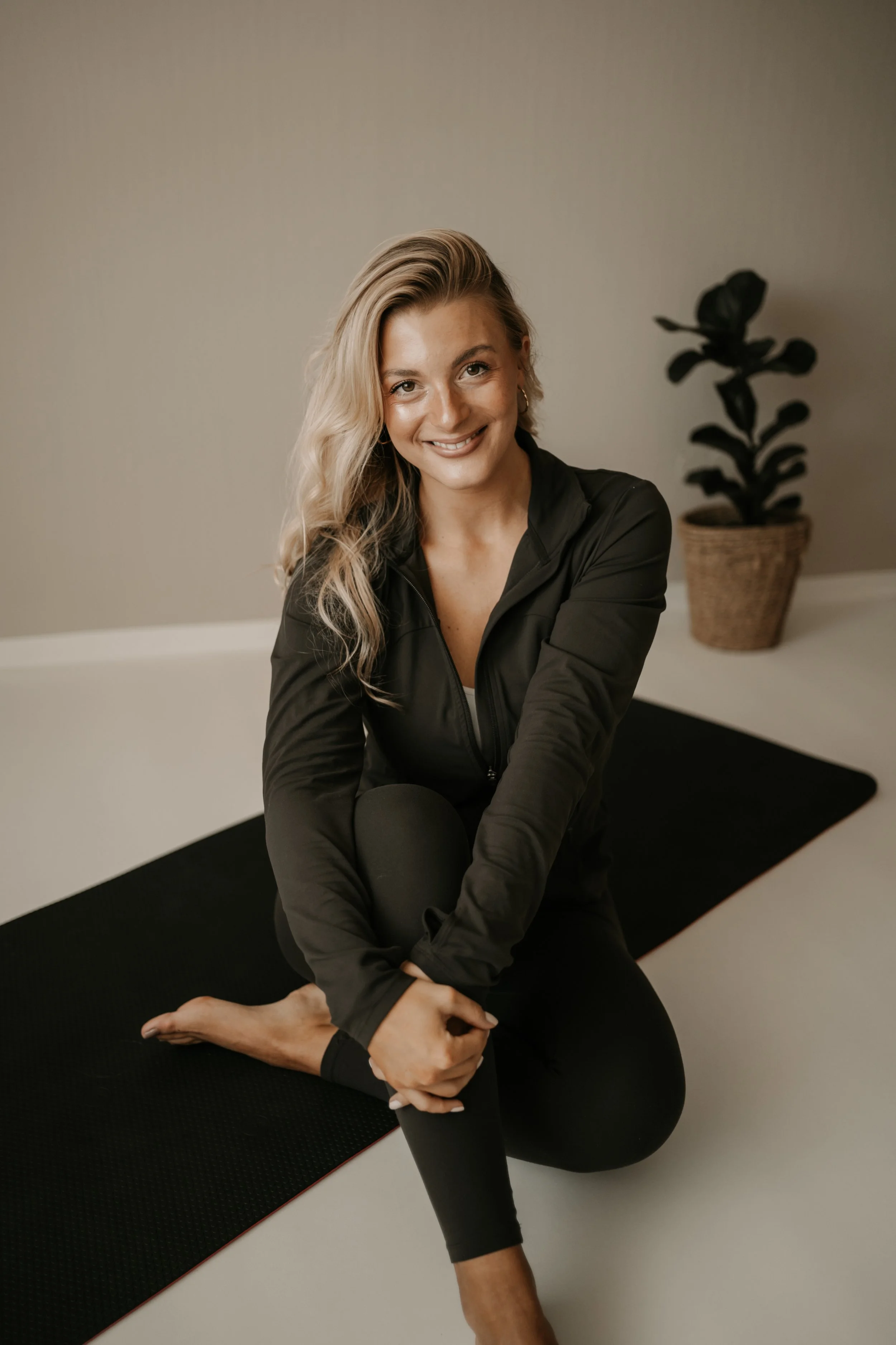 A woman doing yoga on a black mat in a minimal room with a potted plant in the background.