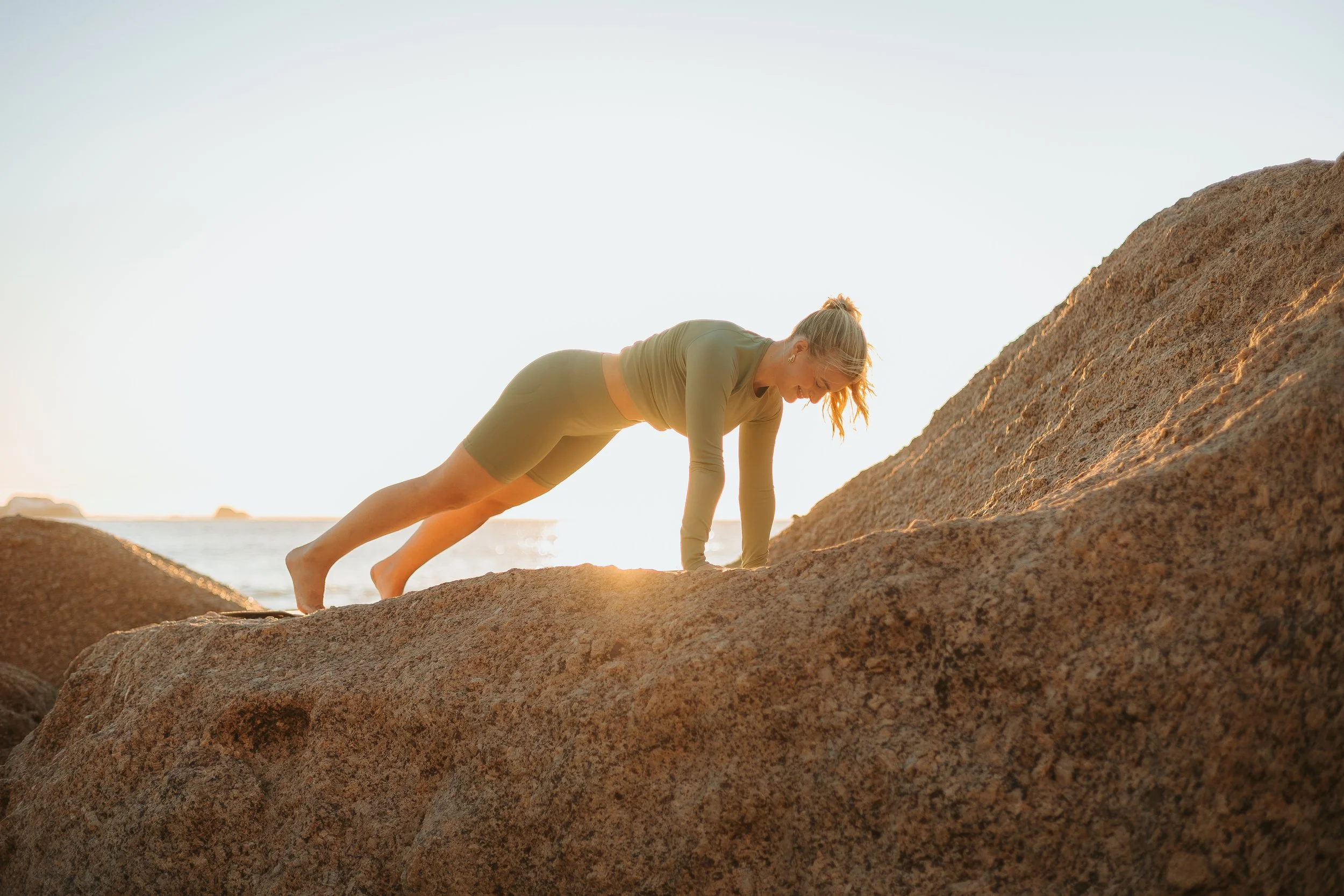 Woman performing push-ups on rocks at sunrise or sunset by the beach.