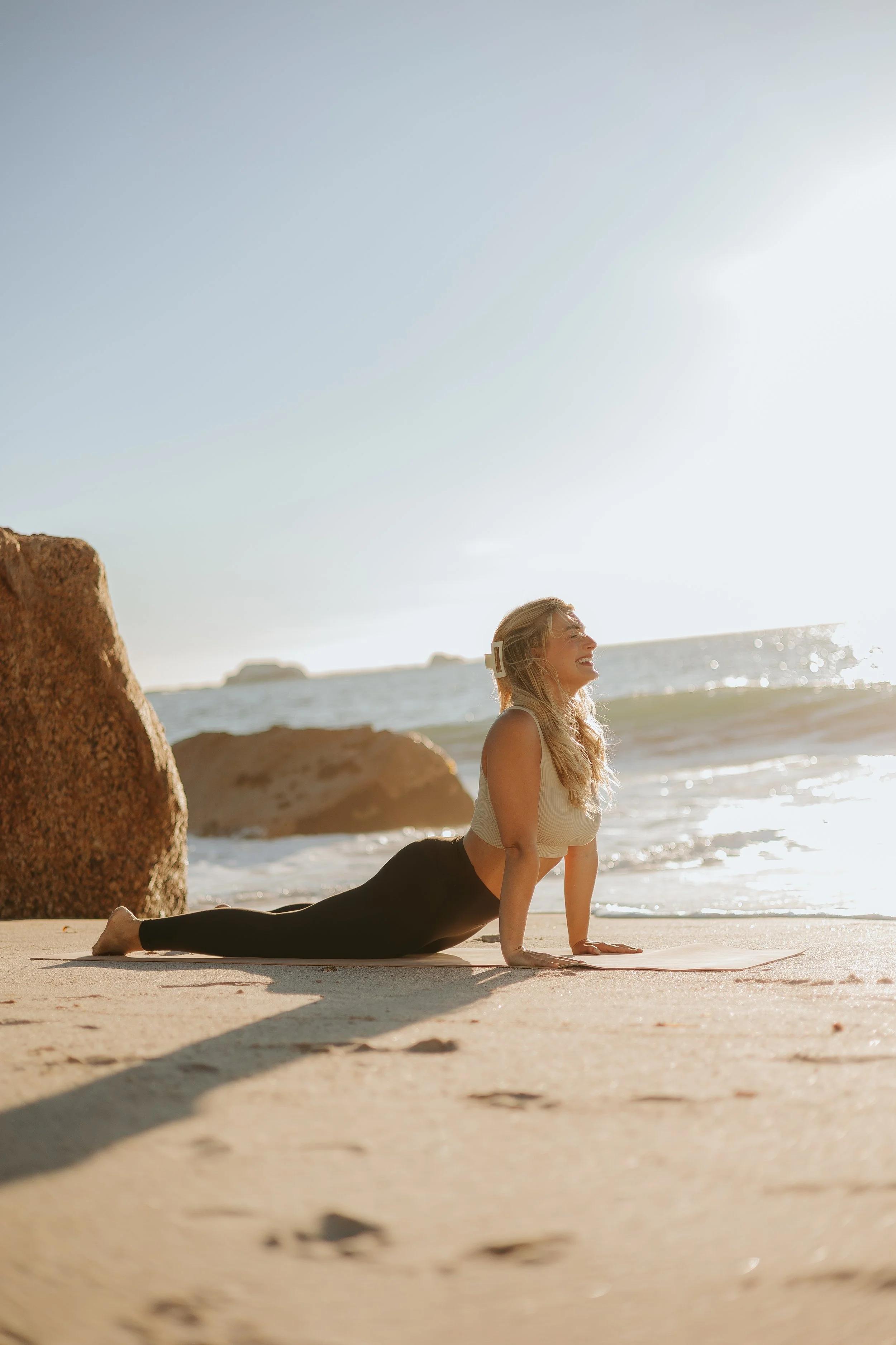 A woman practicing yoga on the beach at sunrise, performing upward-facing dog pose with the ocean waves in the background.