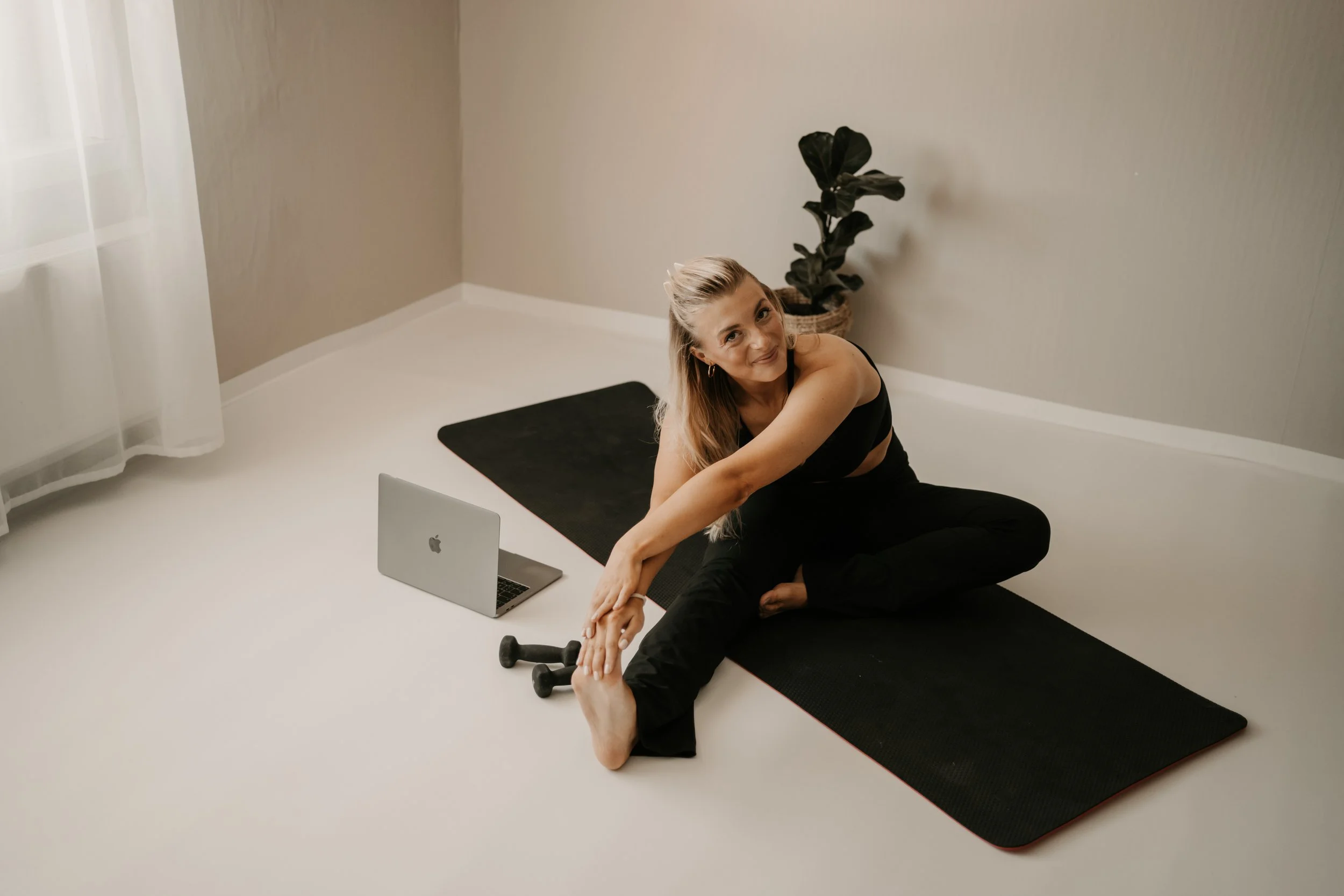 A woman doing a seated yoga stretch on a black mat in a room with beige walls and a white floor, with a laptop, dumbbells, and a potted plant nearby.