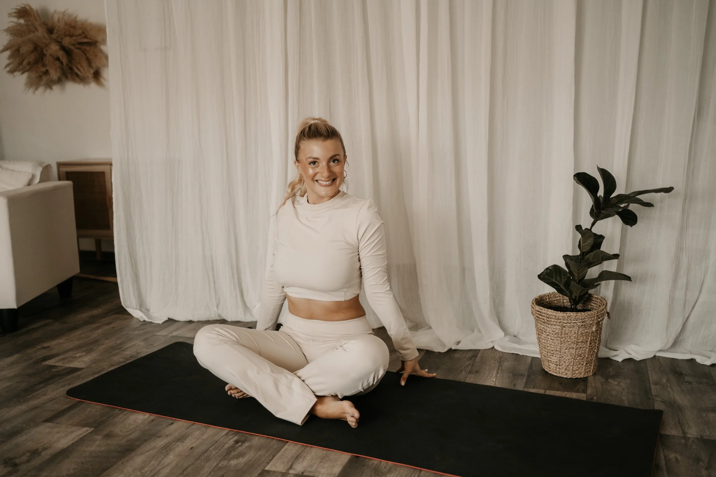 A woman with blonde hair in a ponytail sitting cross-legged on a black yoga mat inside a room with white curtains and wooden flooring, smiling at the camera, with a potted plant nearby.