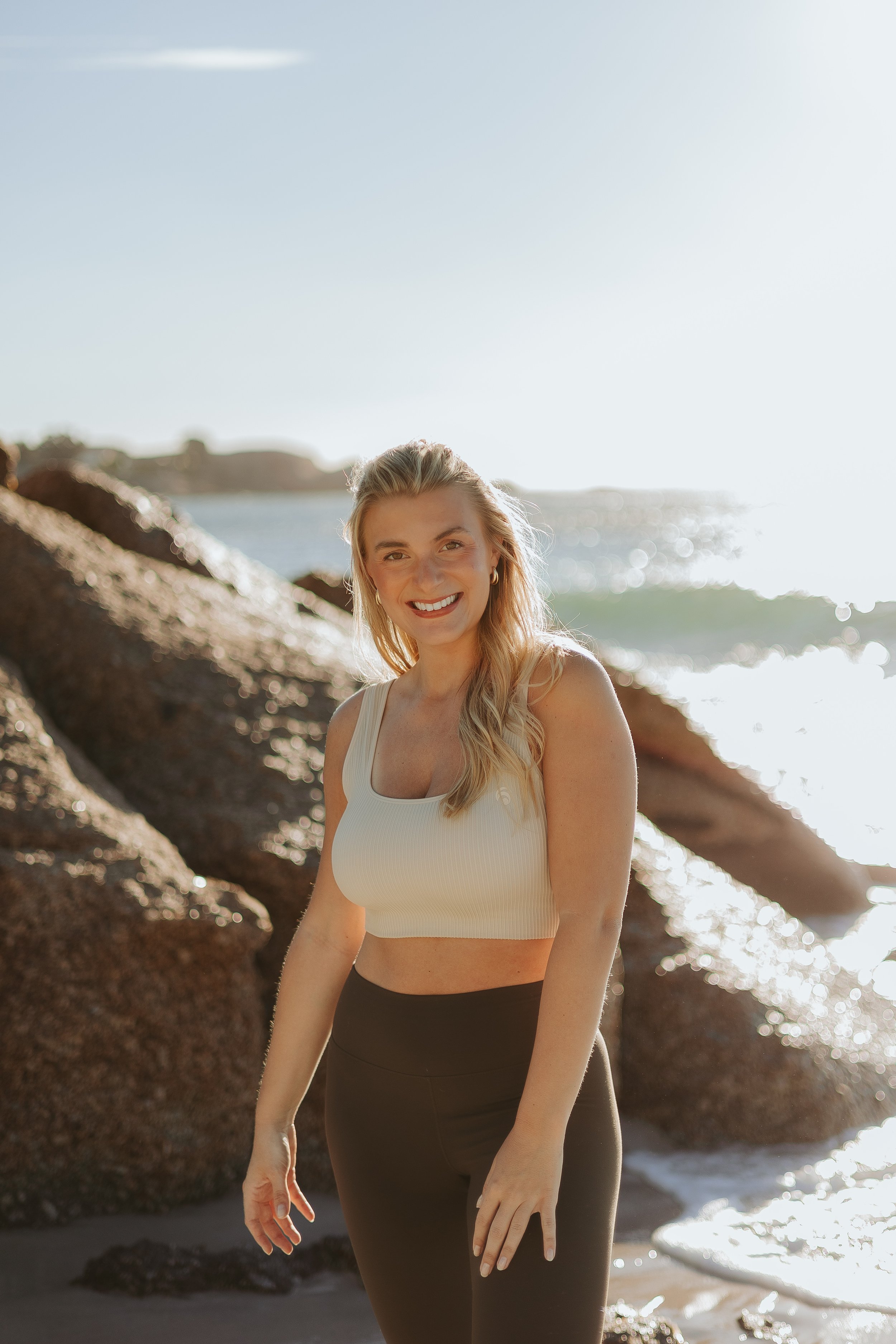 A woman with blonde hair in a beige crop top and black pants standing on a rocky beach near the ocean, smiling with sunlight reflecting on the water in the background.