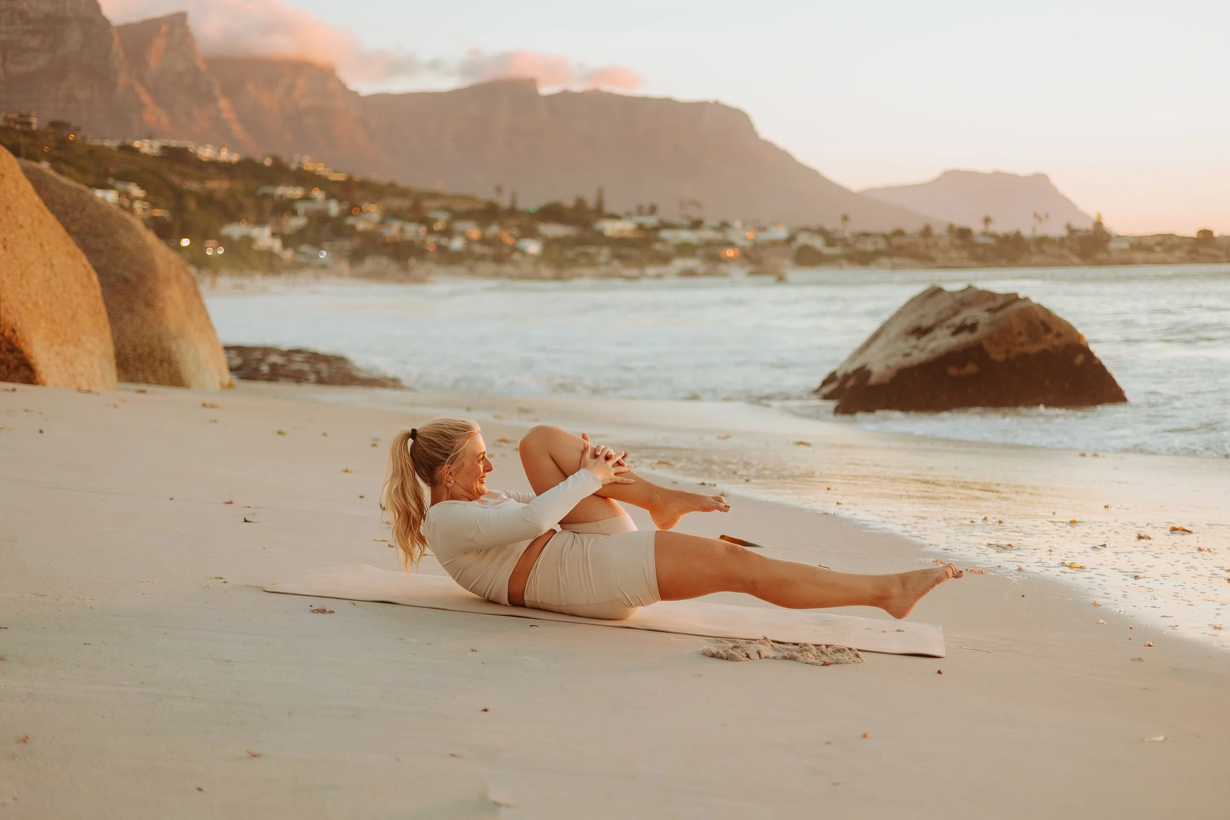 A woman practicing yoga on a beach during sunset, lying on her side on a yoga mat, holding her knee and stretching, with rocks, water, and mountains in the background.