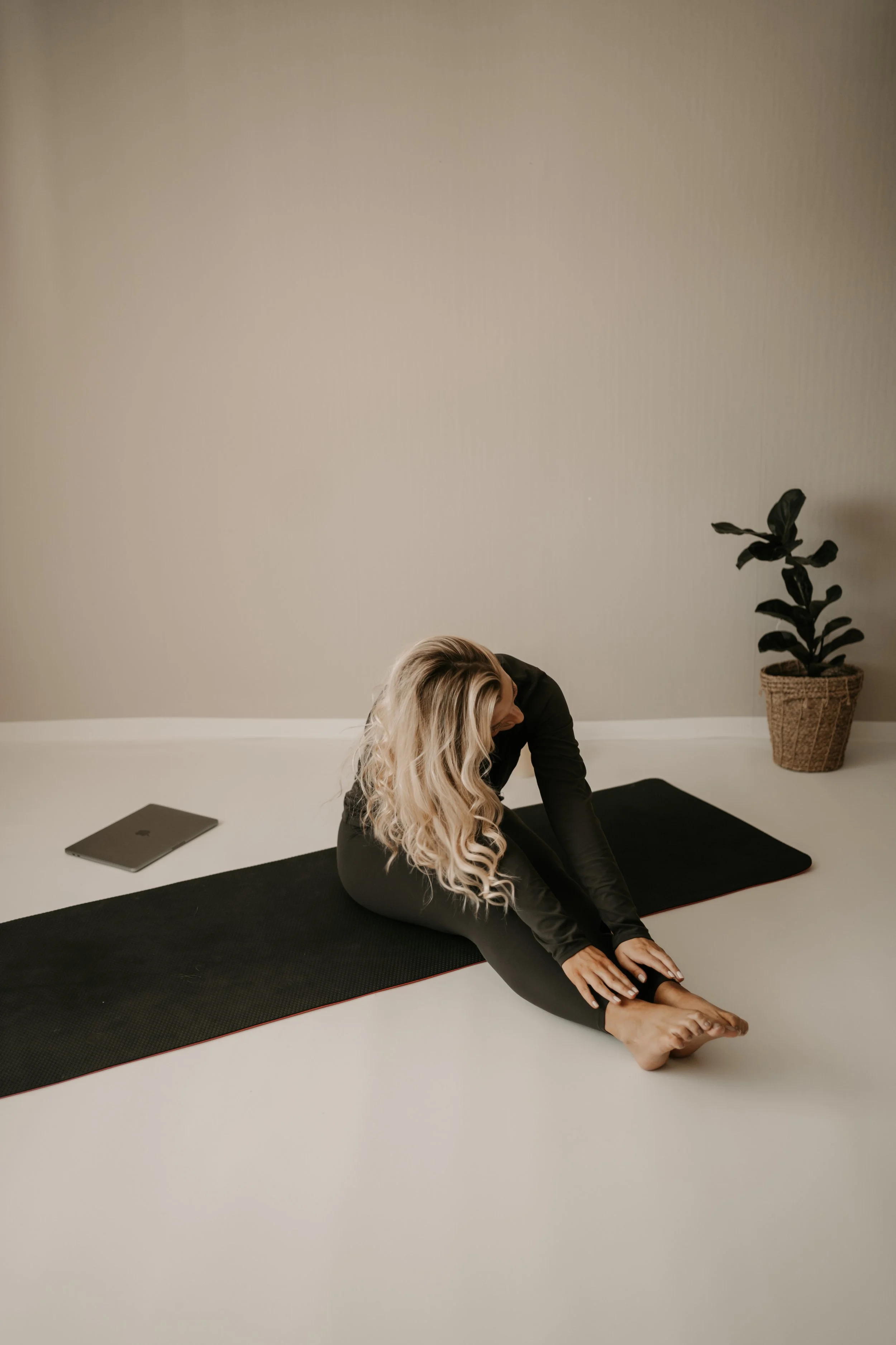 Woman in black activewear practicing seated forward bend yoga stretch on black yoga mat in a minimally decorated room with a potted plant, closed laptop, and blank wall.
