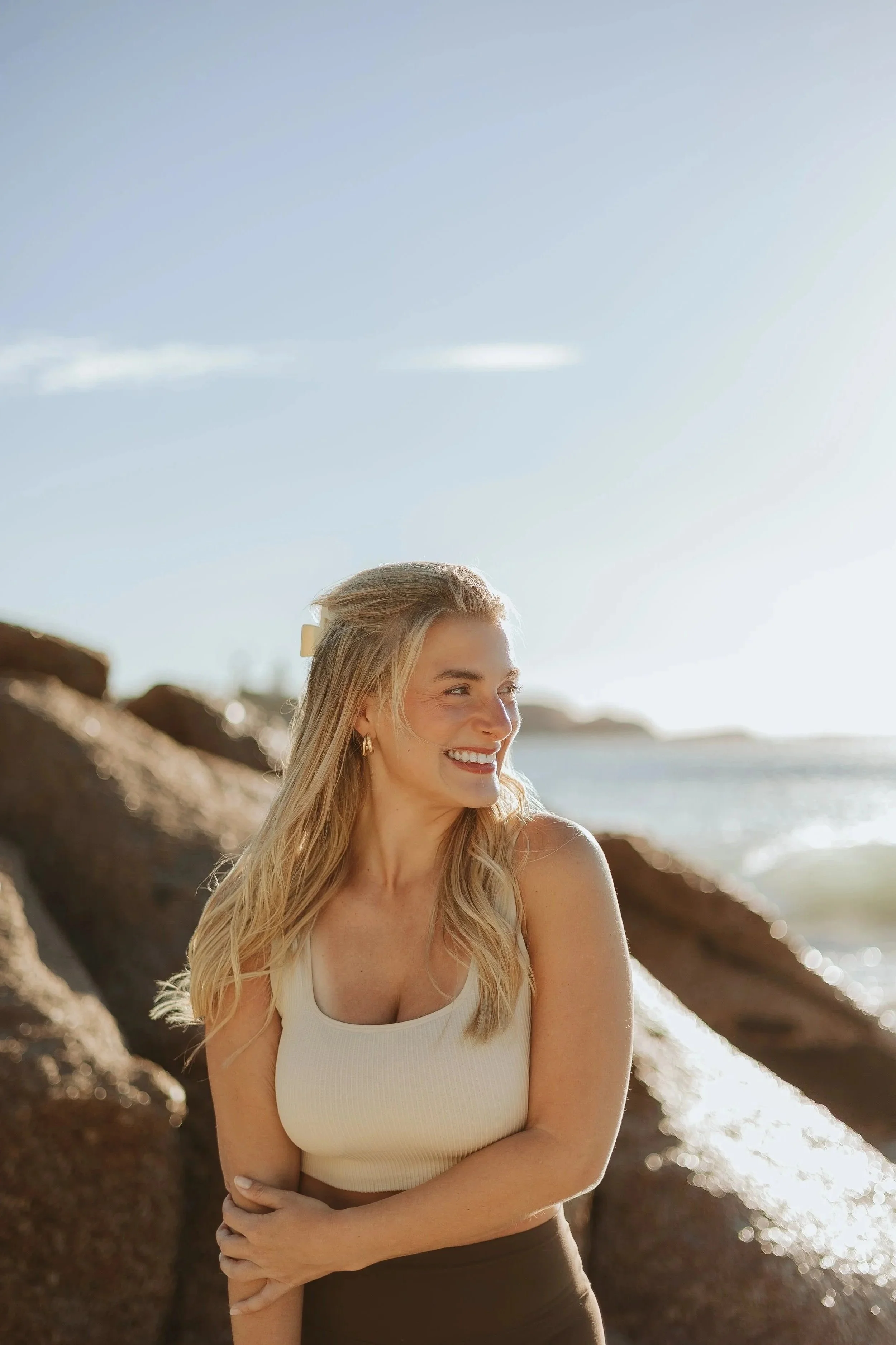 A woman with blonde hair wearing a white tank top and black pants standing on a rocky beach, smiling and looking to her left with the ocean in the background under a clear blue sky.