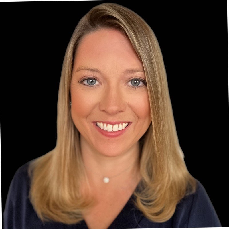 Close-up portrait of a smiling woman with blonde hair and blue eyes, wearing a dark top and a small necklace, against a black background.