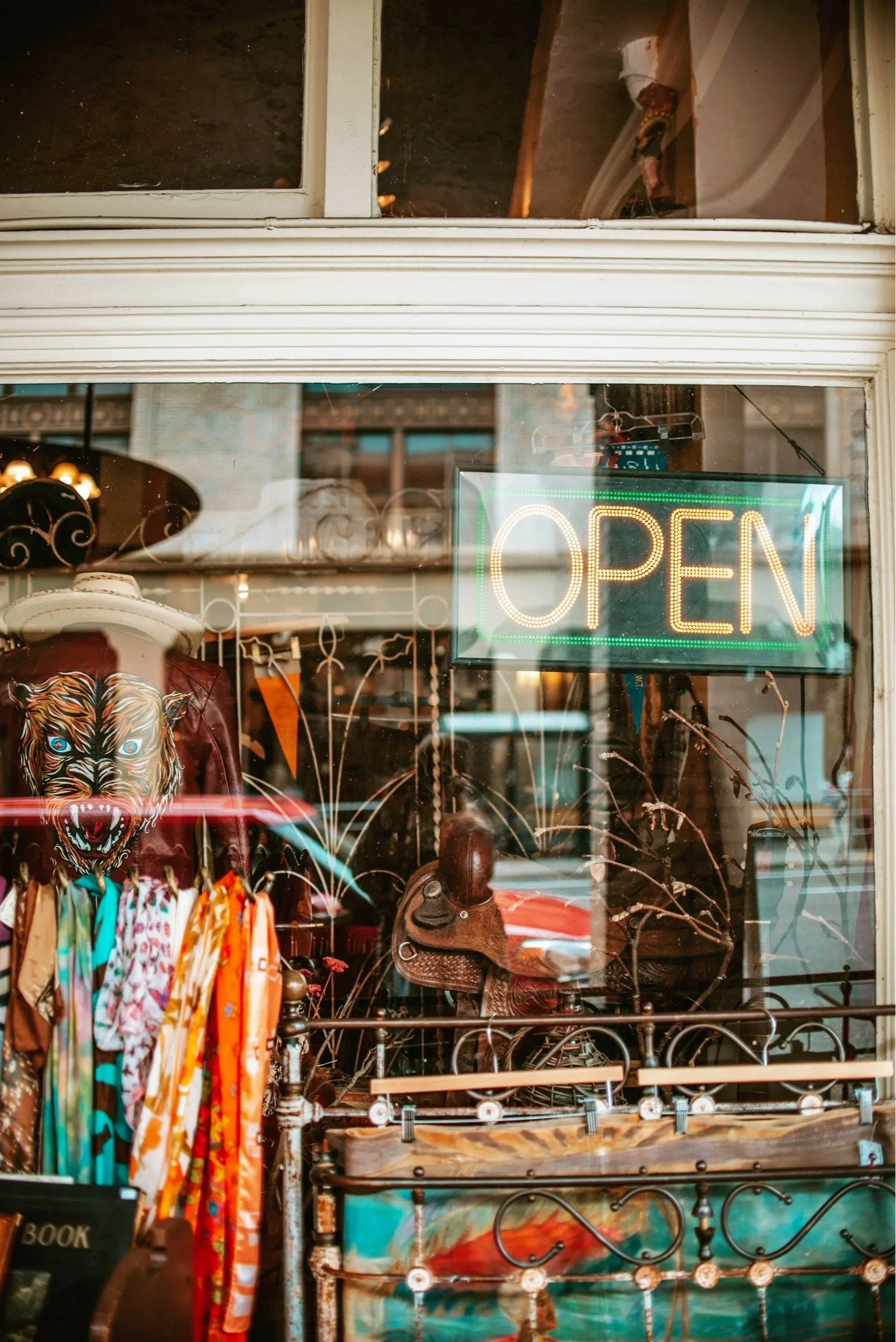 View through a shop window featuring a neon 'Open' sign, a display with a jacket with a tiger face mask, scarves, a cowboy hat, a leather saddle, and a wrought iron and glass decor with branches inside the store.