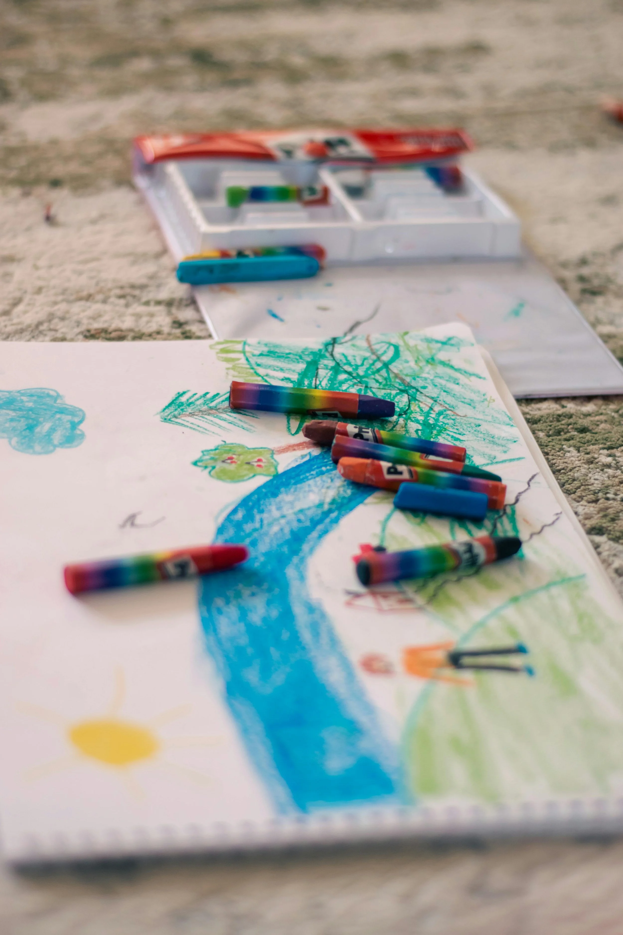 Colorful drawing of a river, trees, and a sun on a white paper, with rainbow-colored crayons scattered on top.