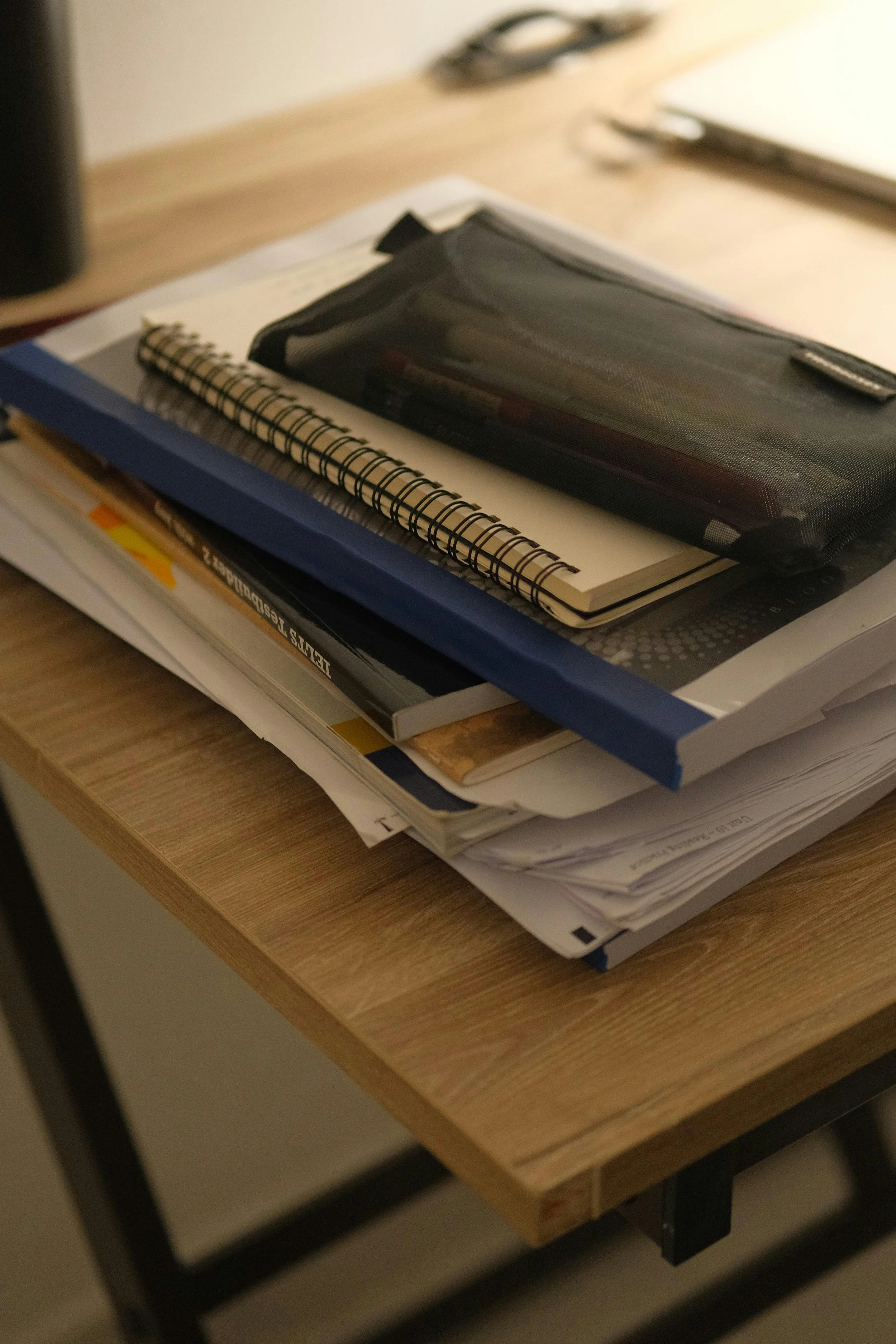A wooden desk with a stack of documents, notebooks, a blue folder, and a black zippered pencil case on top.