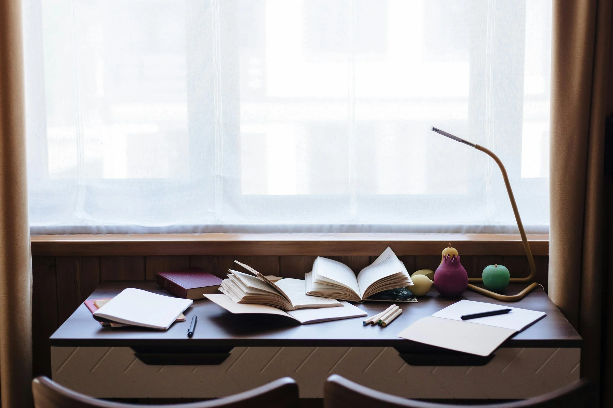 Study desk with open books, notebooks, pens, and decorative apple and egg shaped objects, placed in front of a window with curtains.
