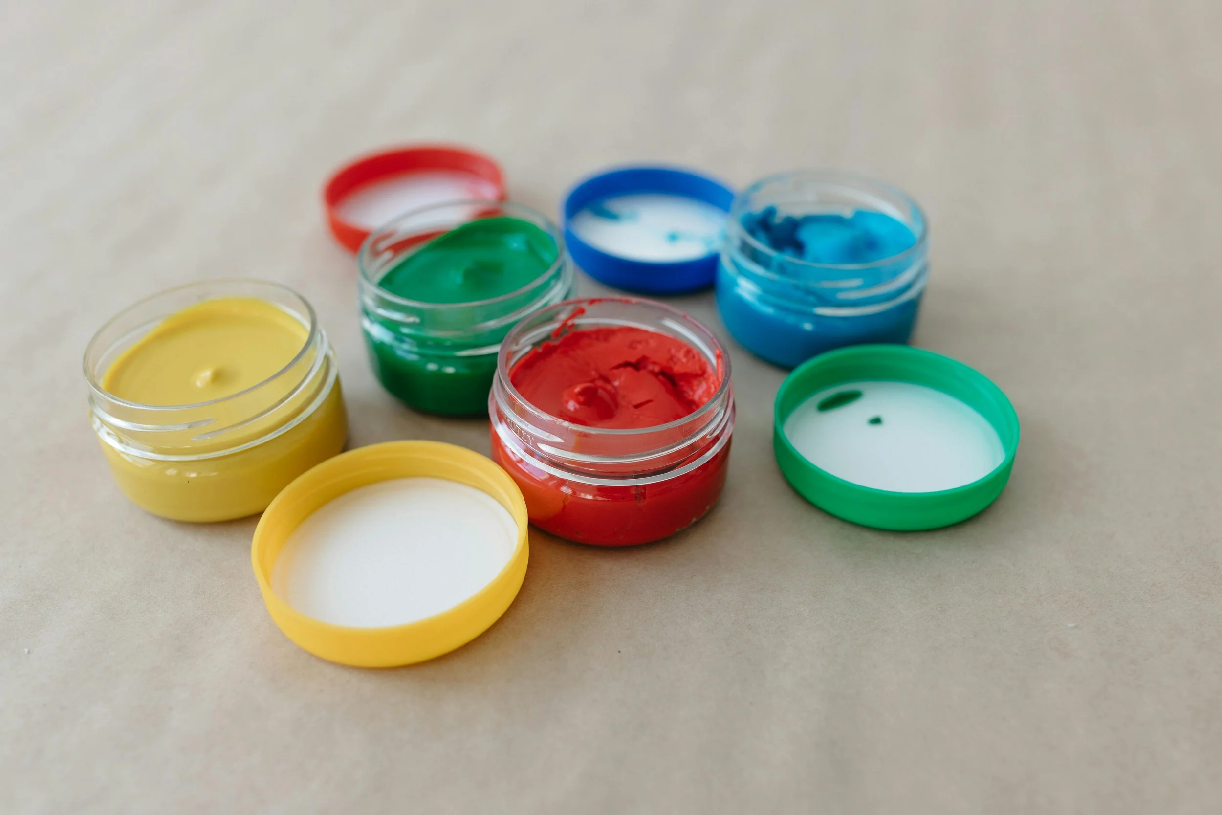Open jars of colorful paint in yellow, red, green, blue, and white on a beige surface.