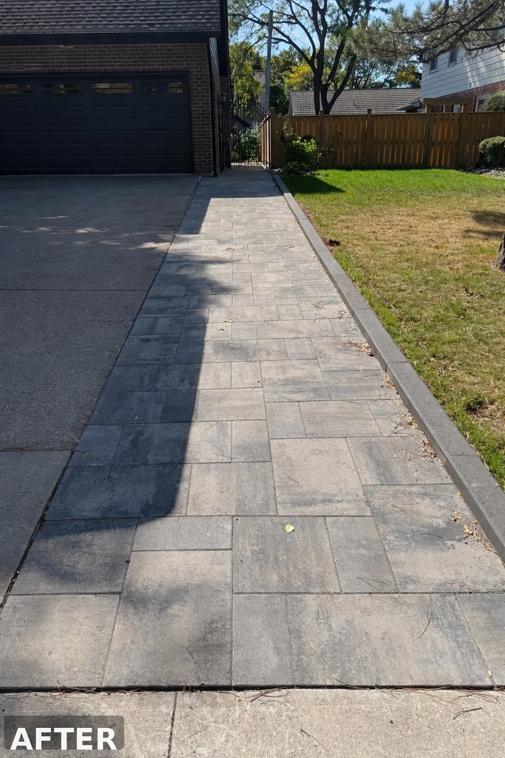 Newly paved concrete sidewalk with a clean edge, adjacent to well-maintained grass and a dark brick house in the background, under clear sky with tall trees.