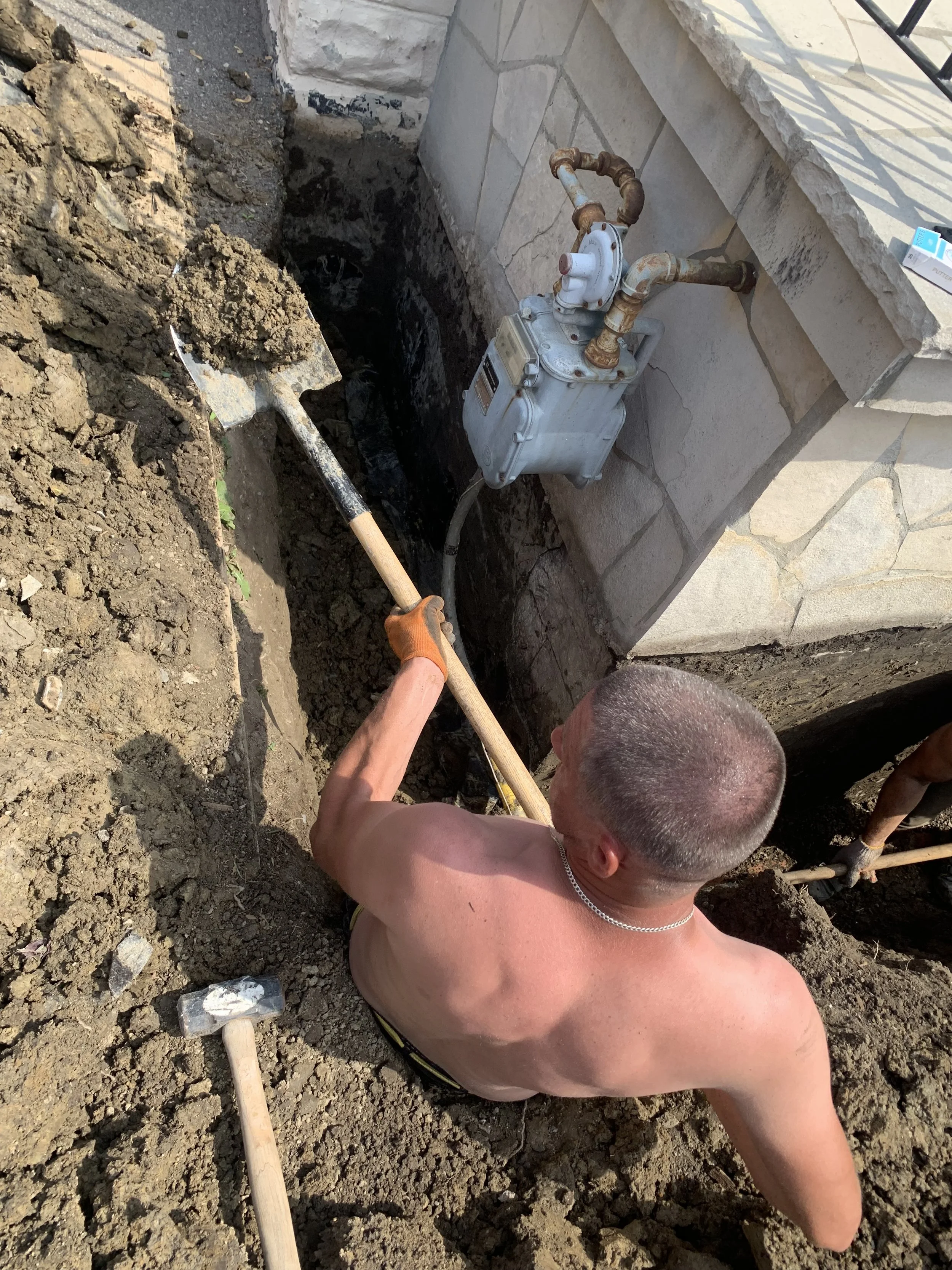 A man with a shaved head and no shirt working in a dirt trench, using a shovel, next to a house with a stone exterior and a gas meter.
