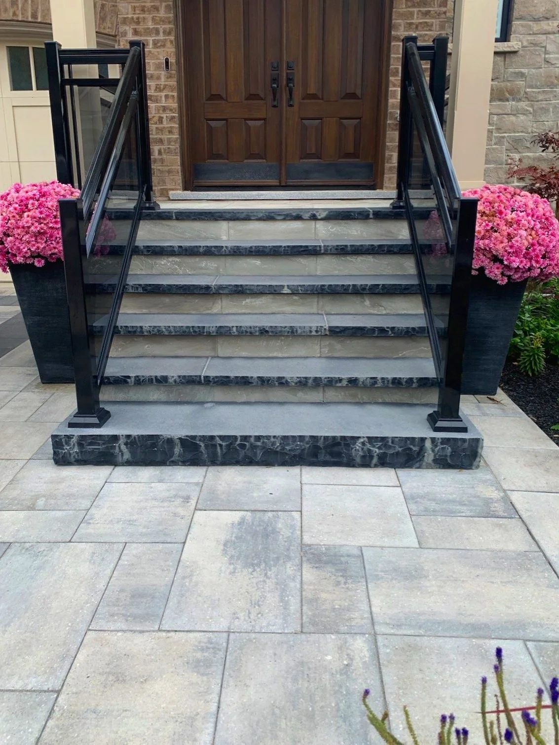 Front porch with marble steps, black metal railings, and pink potted flowers on both sides. A wooden door is at the top of the stairs.