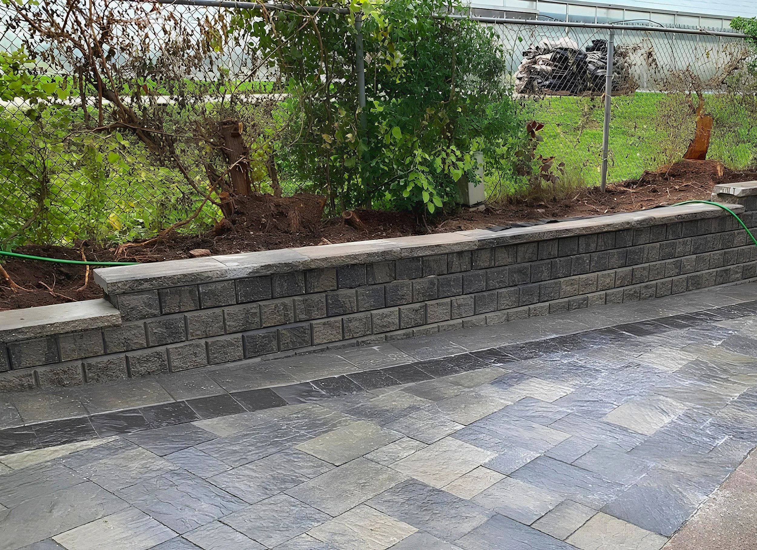 A newly constructed brick retaining wall with dark gray bricks separates a garden bed with plants and soil from a paved patio area with gray stone tiles.