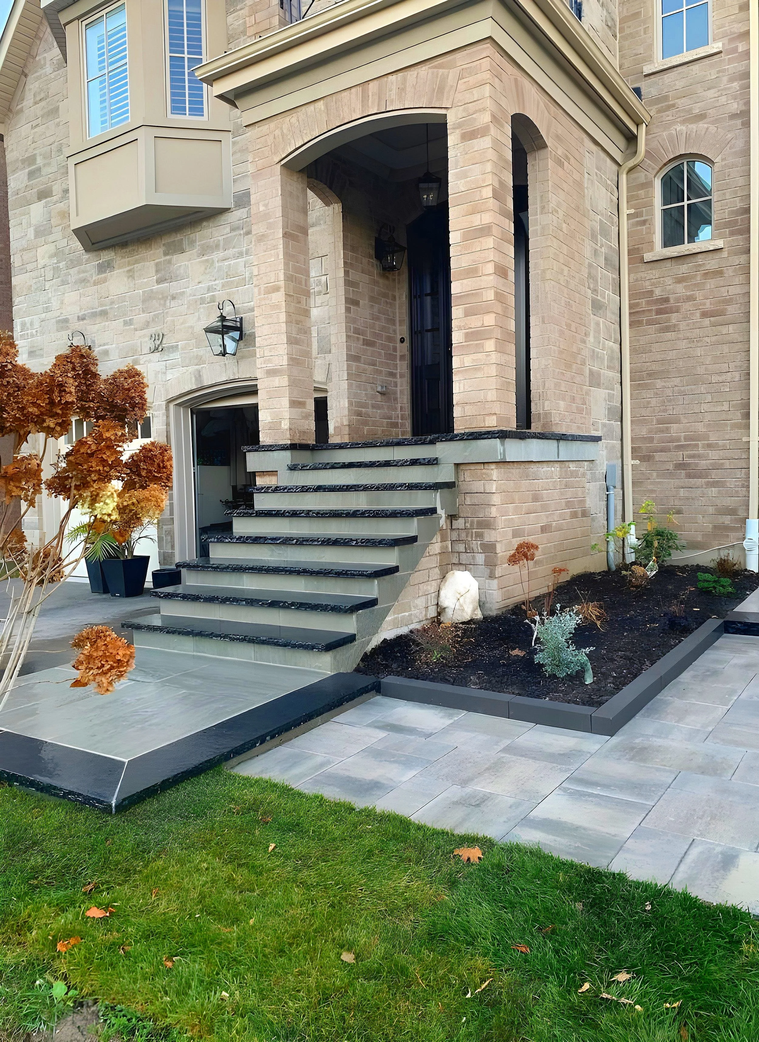 Exterior view of a modern brick house with a front porch, black front door, and stairs leading up to the entrance. There is a small garden with plants and mulch, a potted tree, and a mix of stone and brick paving on the ground.