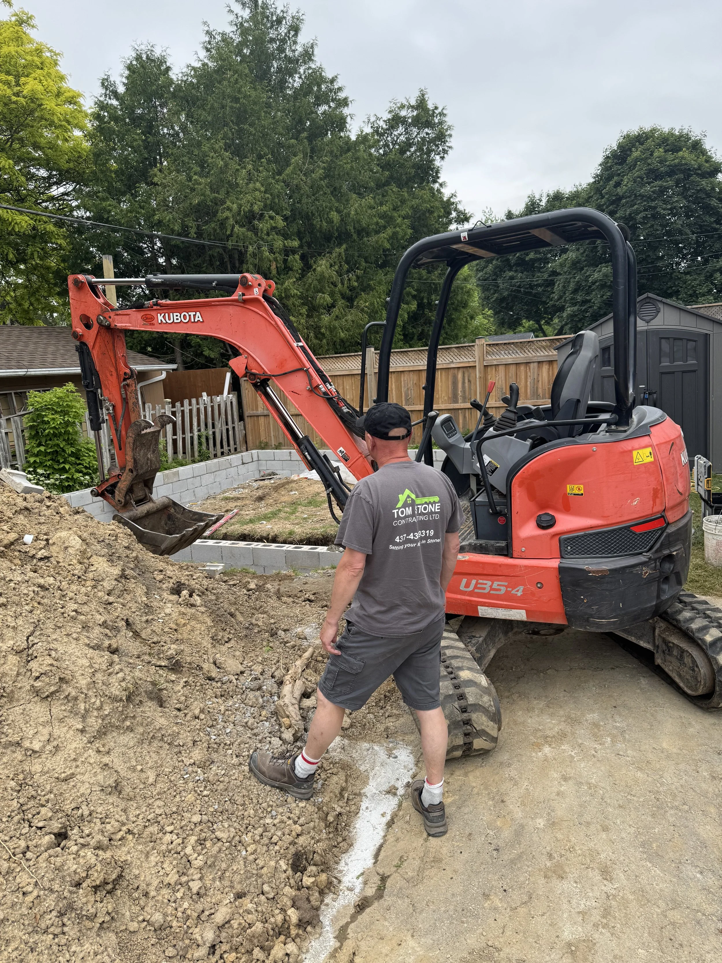 A construction worker in casual clothes and a cap operating a small orange Kubota excavator digging a trench in a backyard, with a man standing near the pile of dirt.