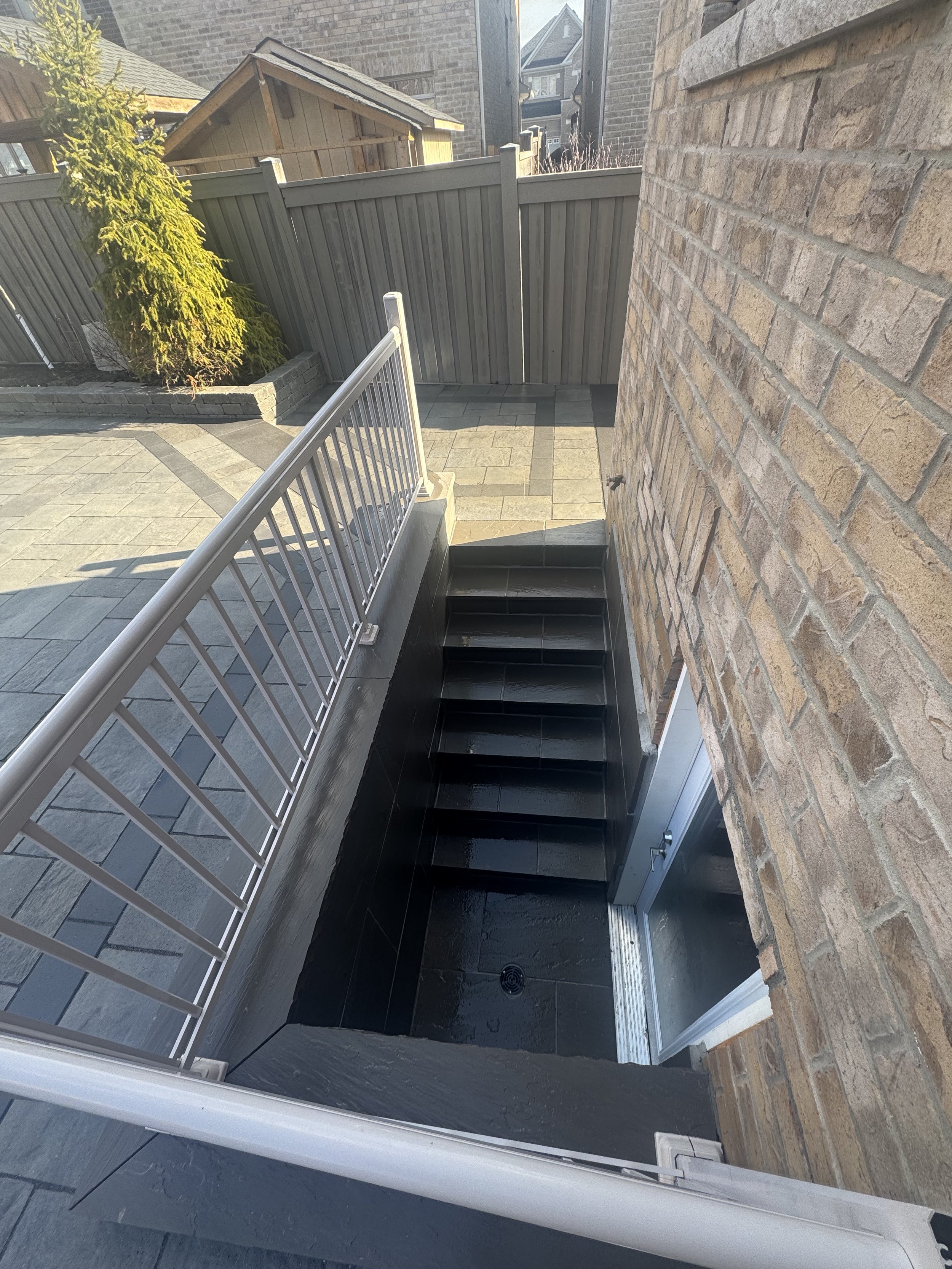 Outdoor patio with black stairs leading down to a door, brick wall on the right, gray wood fence in background, and a small green tree in a raised brick planter on the left.