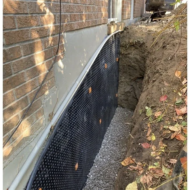 Drainage or waterproofing installation along the side of a brick house with a trench and black waterproof membrane.