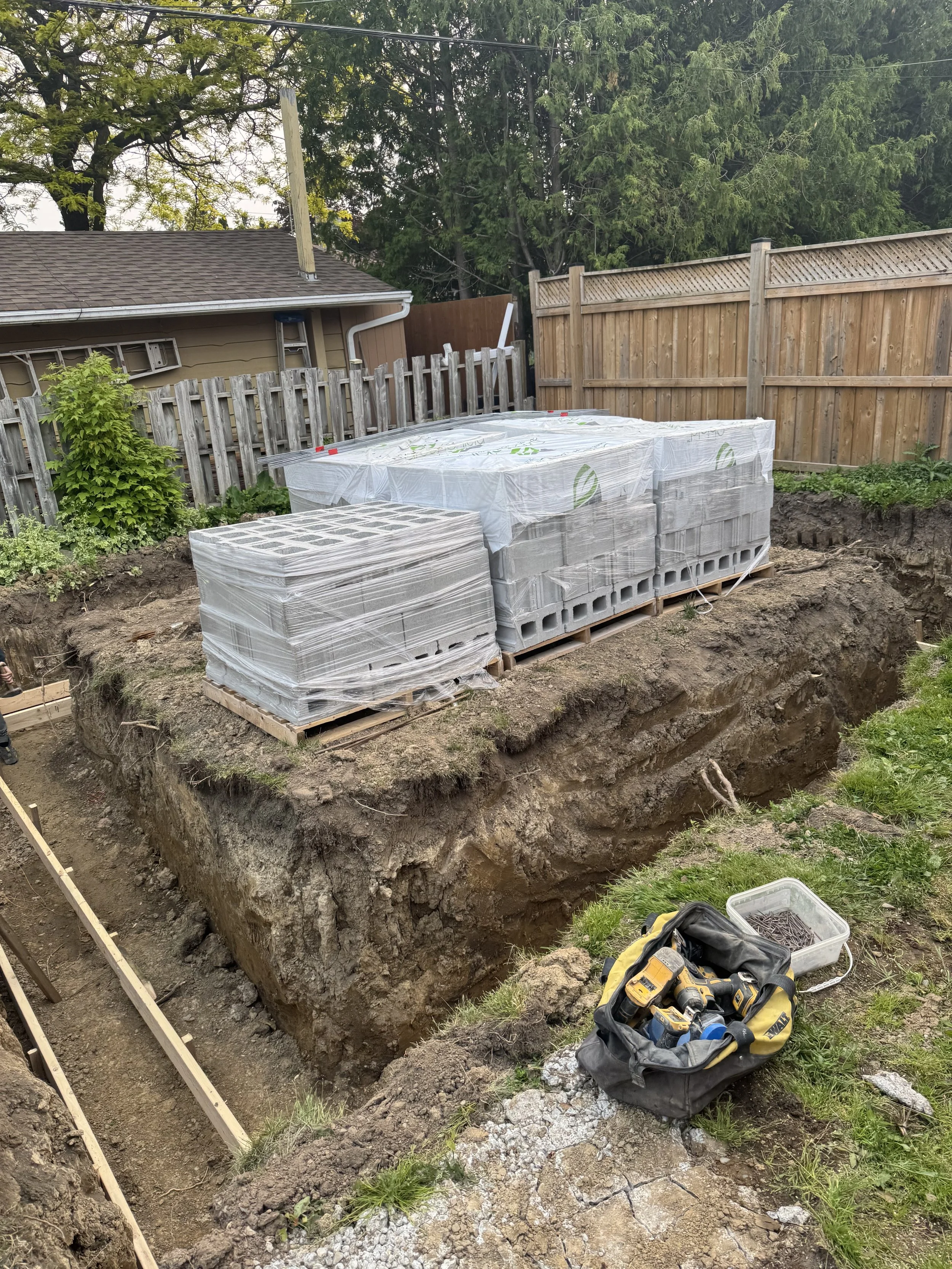 Construction site with an excavated hole, pallets of concrete blocks covered with plastic, a tool bag with power tools, and a container of nails in a backyard with a wooden fence and trees.