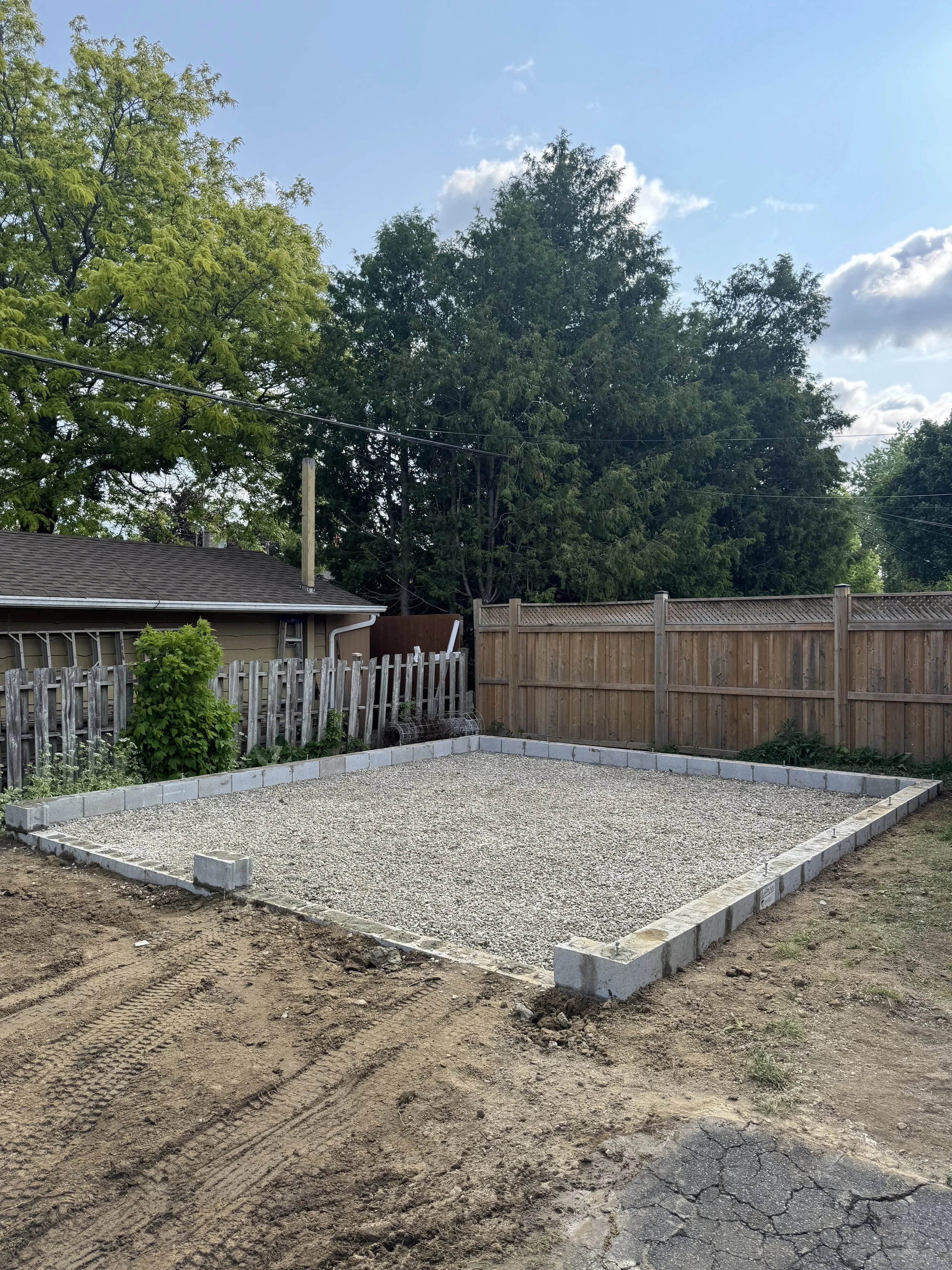 A small backyard fenced with wood, with a gravel-filled area outlined by concrete blocks, and some plants along the left fence. The sky is partly cloudy and there are trees in the background.