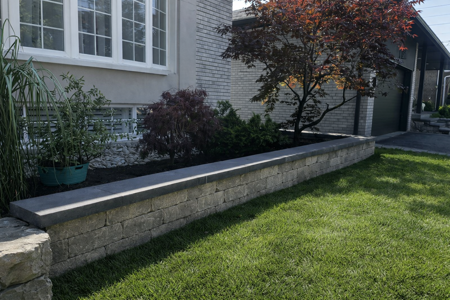 A landscaped front yard with a brick wall, containing green grass, trees, and bushes, alongside a house with a white exterior and large windows.