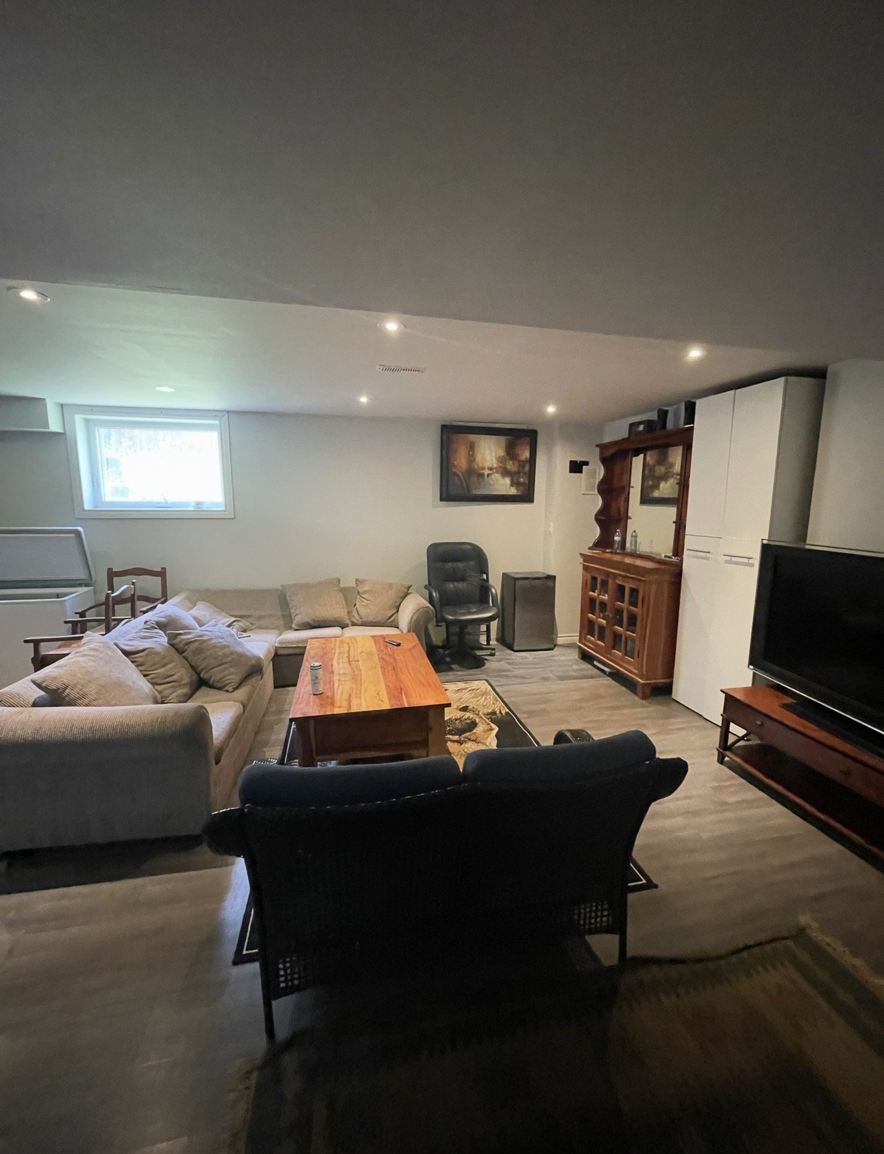 A basement living room with beige sofas, a wooden coffee table, a black reclining chair, a TV on a stand, a wooden cabinet, and framed artwork on the wall, with a small window and wood flooring.