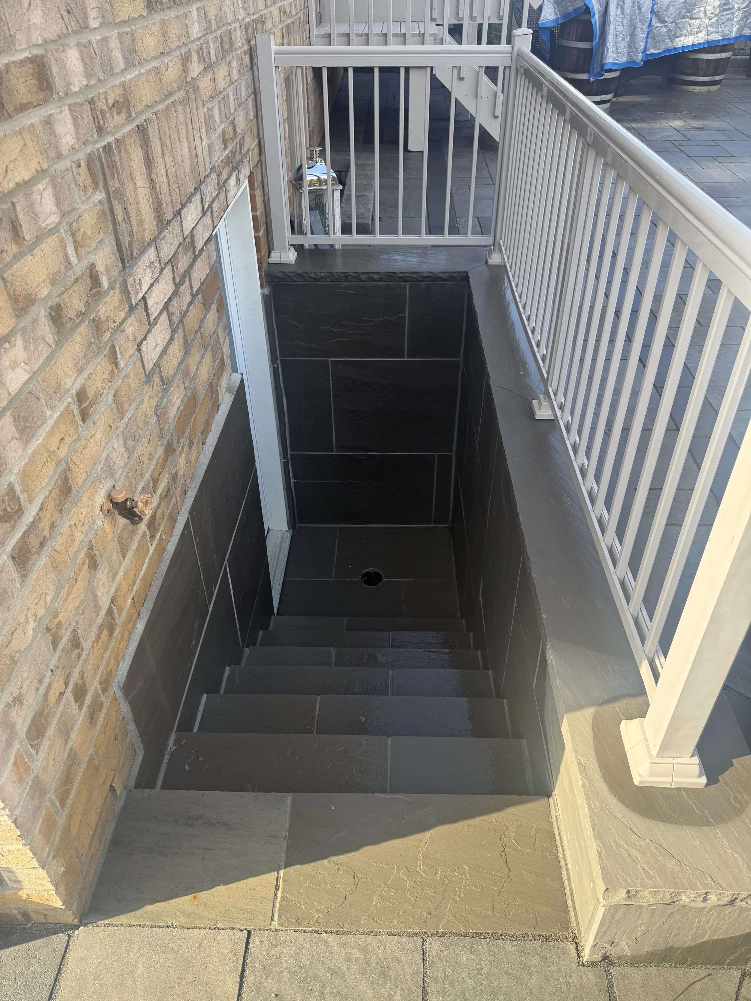 A small outdoor staircase leading down to a sloped paver pathway, with black tiles on the walls and steps, surrounded by a brick wall on one side and a white railing on the other.
