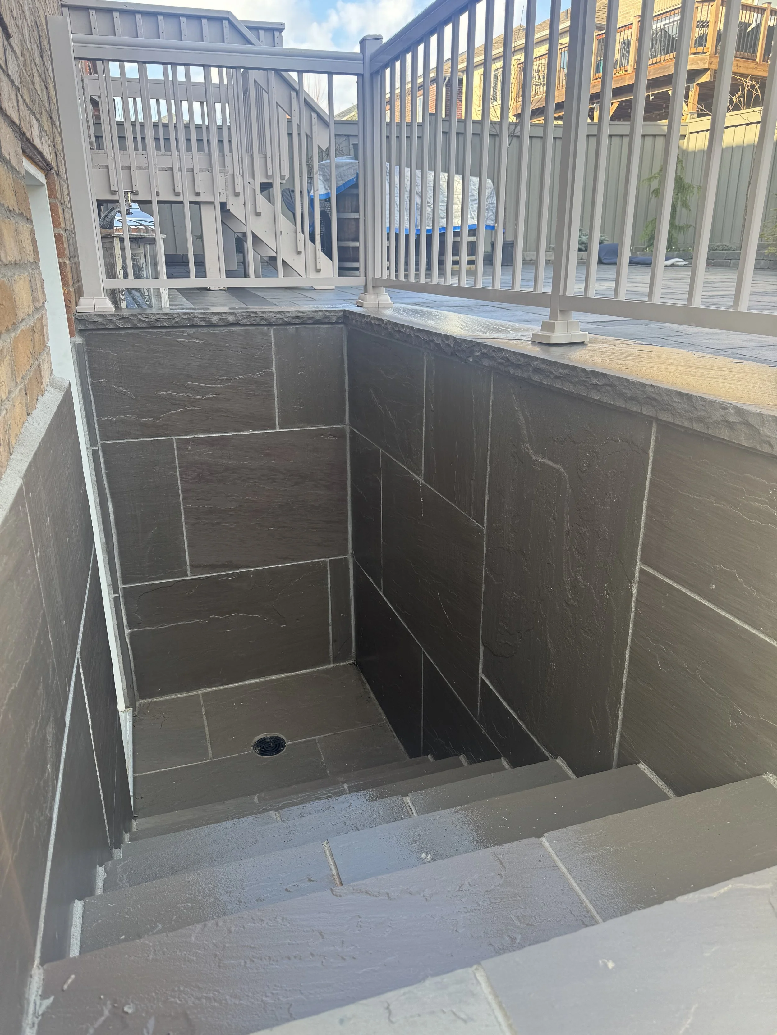 Underground stairway with dark tile walls and steps, leading downward, with a drain at the bottom, surrounded by a concrete patio and a white metal railing.