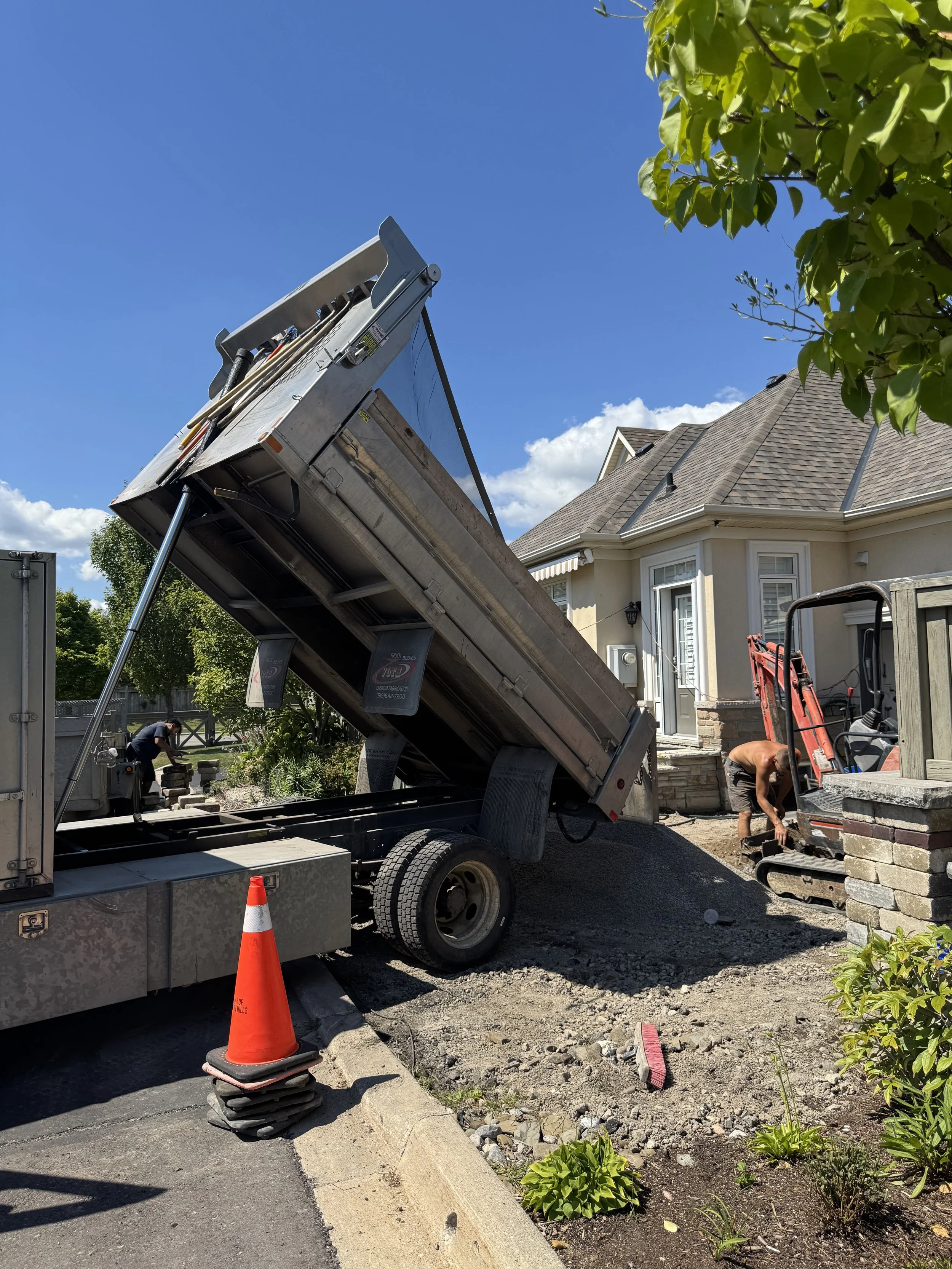 Construction workers are installing a new driveway with a dump truck unloading gravel or soil, a small excavator, a traffic cone, and a house with a beige exterior and brown roof in the background.