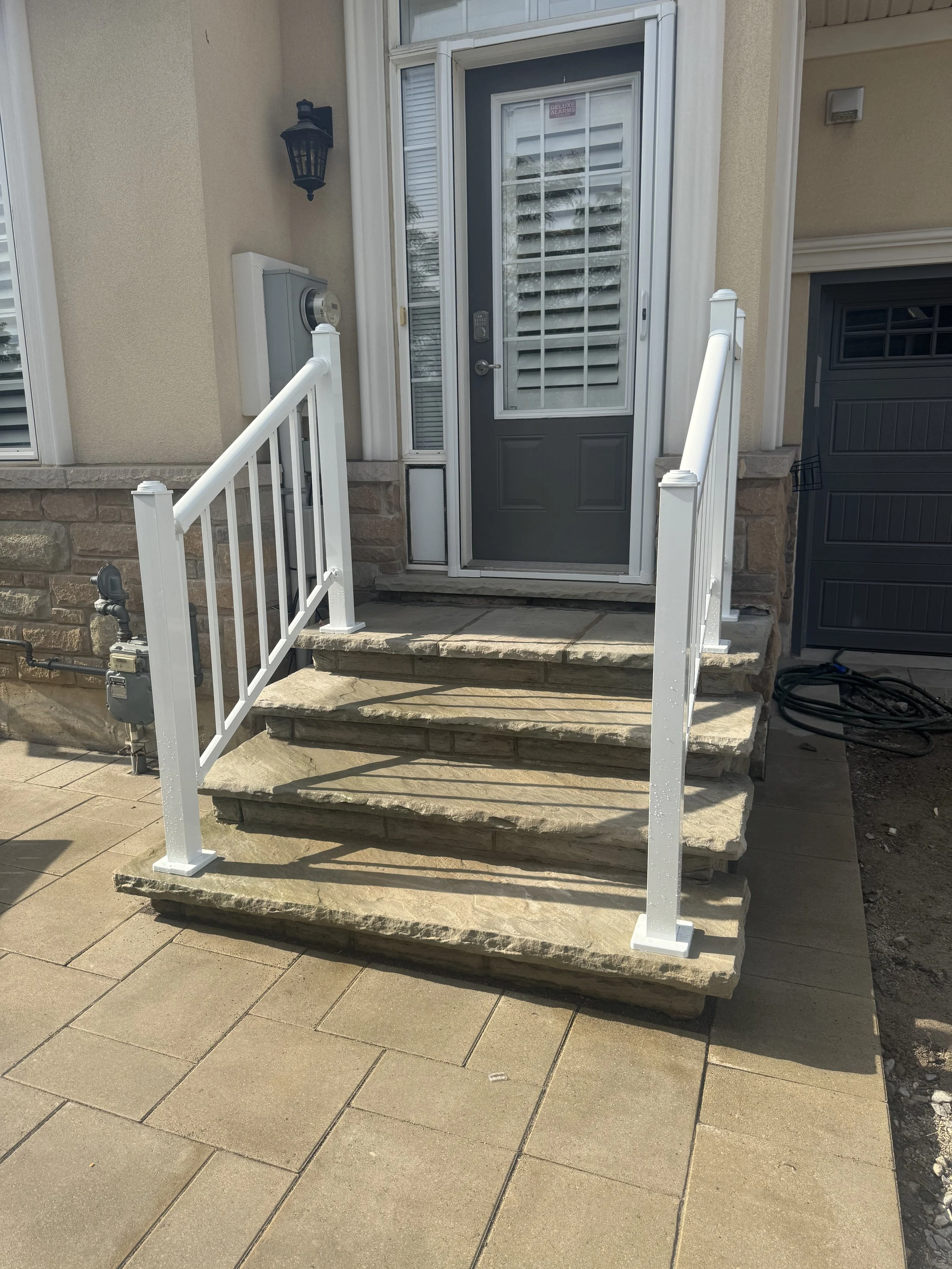 Stone steps with white metal railing leading to a gray front door with glass panel and white blinds, part of a beige house with stone foundation and outdoor light fixture.