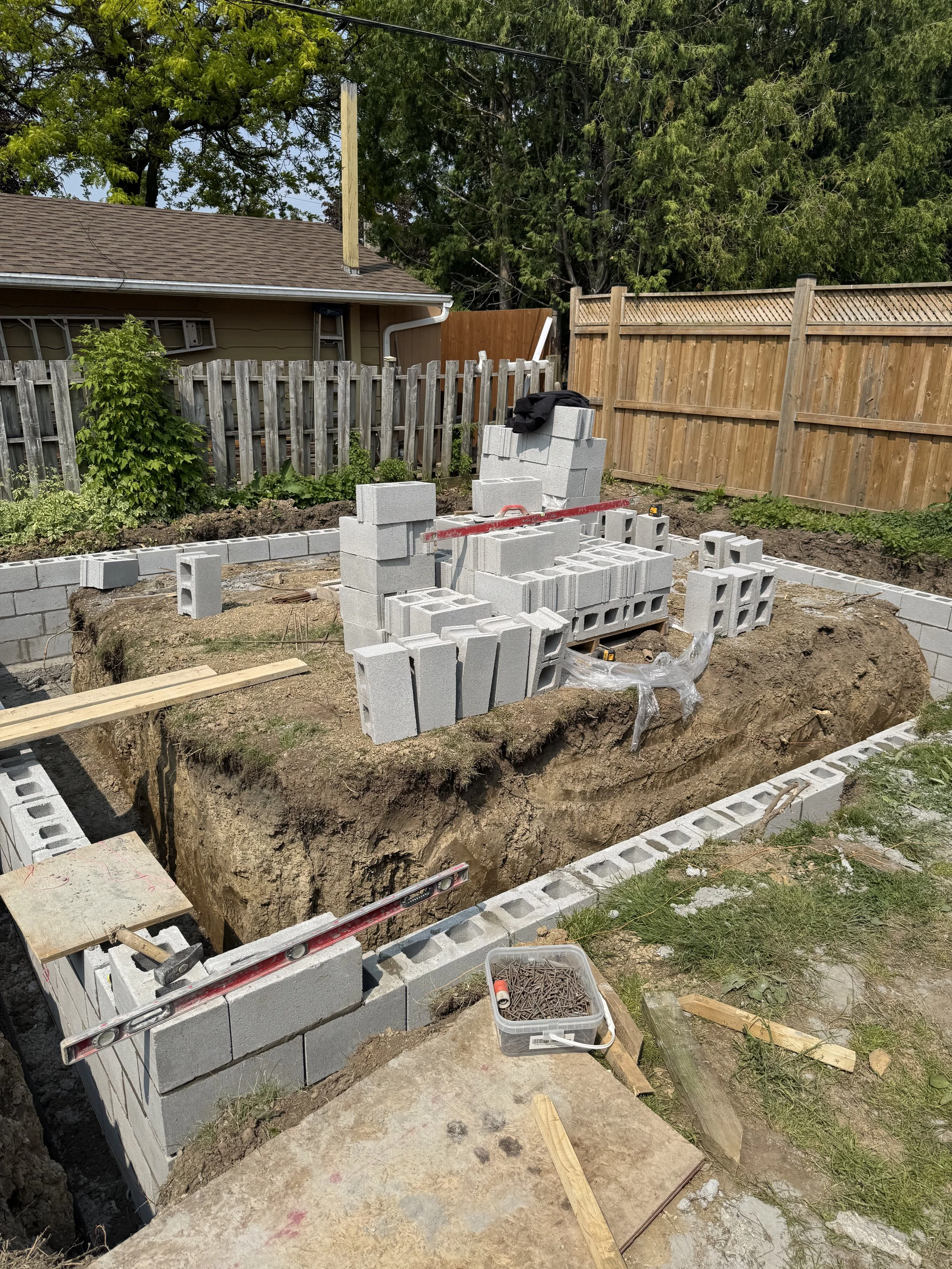 Construction site with a partially built brick or cinder block wall, construction tools, and materials spread around the excavation area, surrounded by a wooden fence and trees.