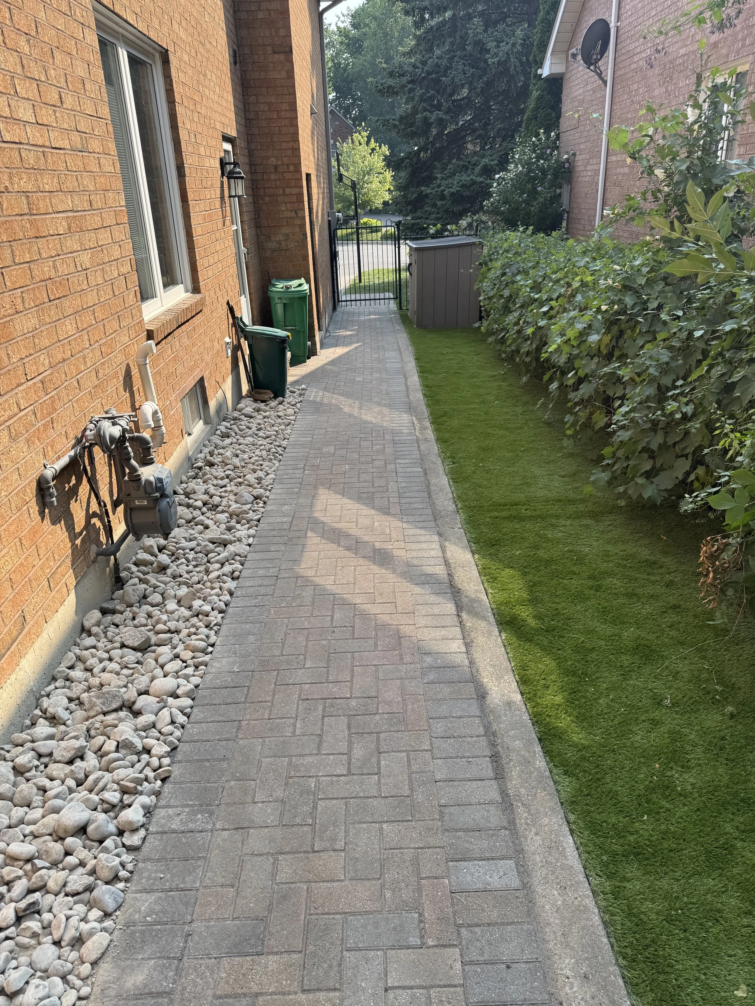 Sidewalk with brick pavers running between a red brick house with two windows and utility meters on the left, and a green lawn with bushes on the right, leading to a black gate and a street in the background.