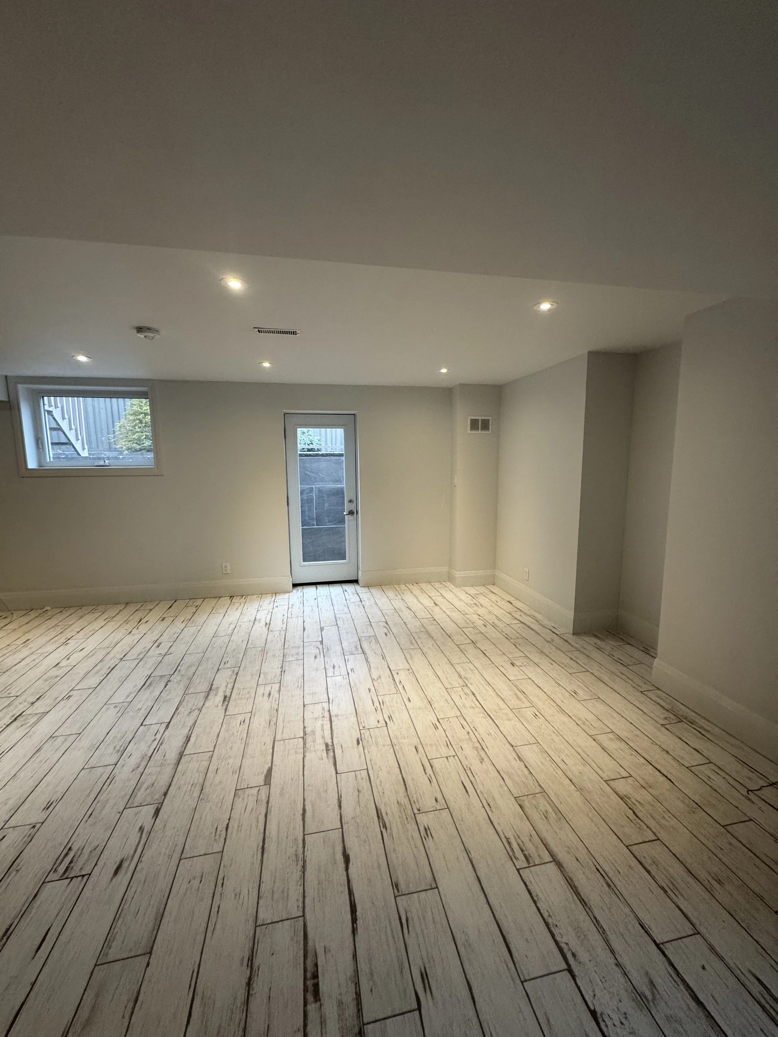 Empty room with hardwood floors, a window, and a door, painted white walls, and recessed ceiling lights.