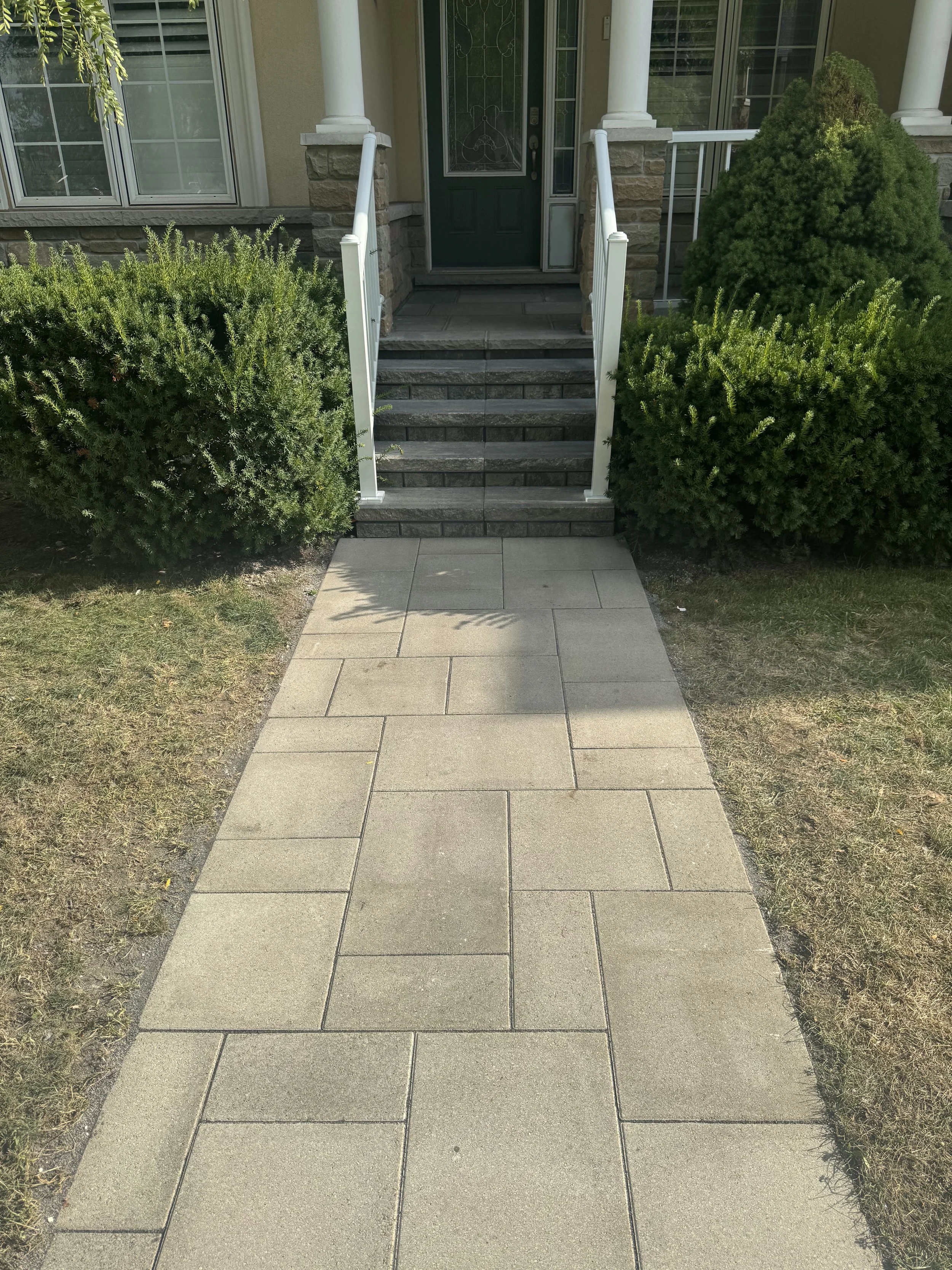 Front entrance of a house with a sidewalk leading to a small staircase, green front door, two bushes on either side of stairs, and a porch with white columns.