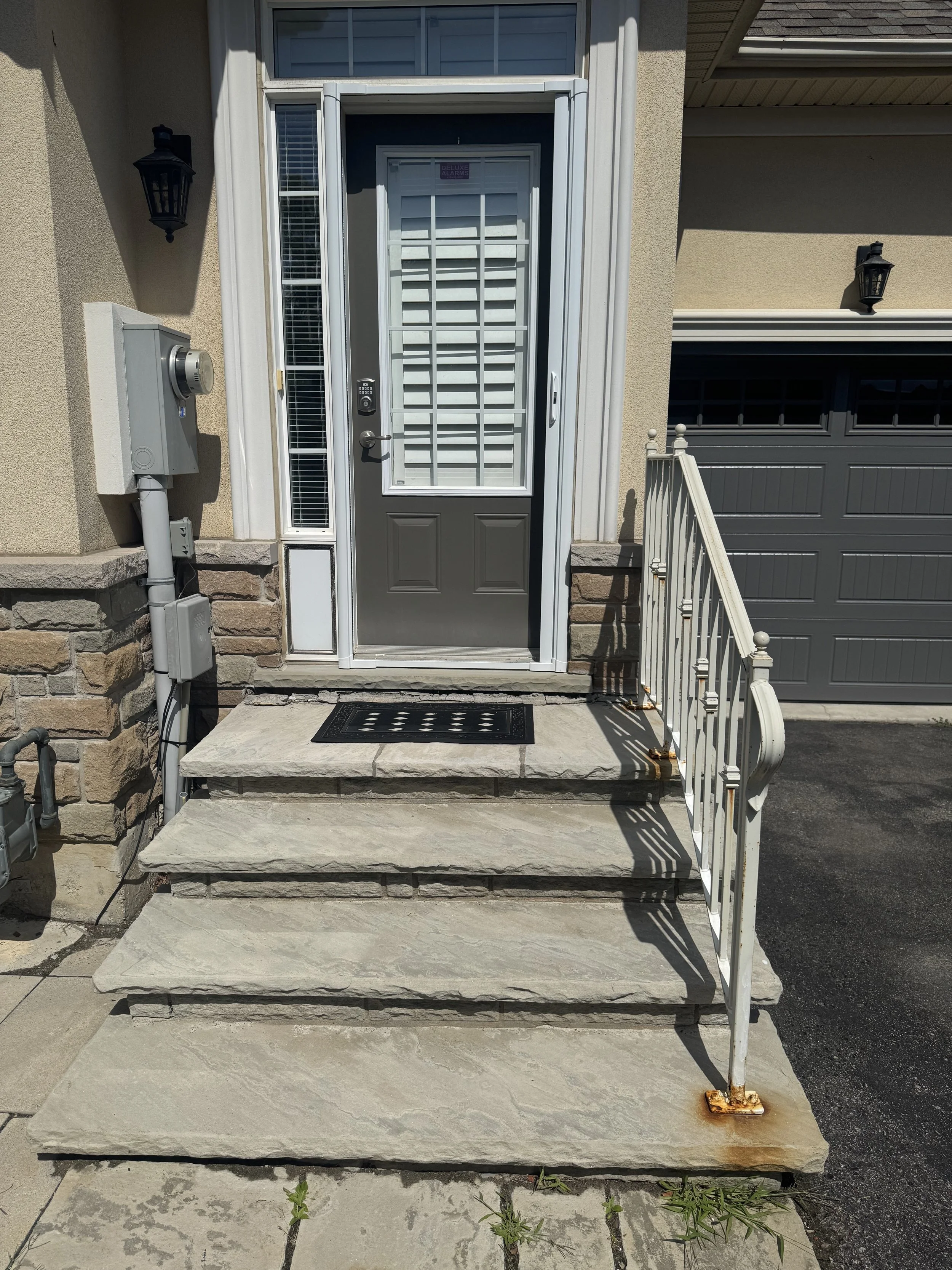 Front entrance with four concrete steps, a gray door with a security screen, black doormat, white metal railing with rust, stone wall accents, light fixtures, and garage with black driveway.