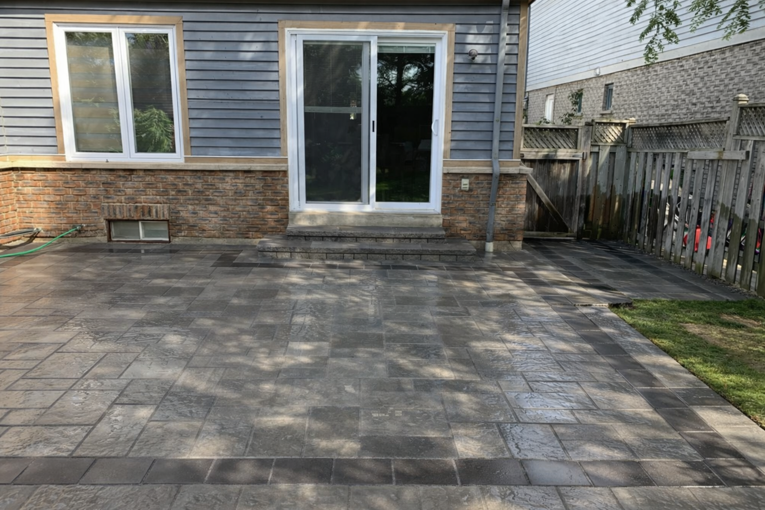 Backyard patio with gray stone tiles, steps leading to sliding glass door, wooden fence on right, house exterior with vinyl siding and brick foundation, small basement window.