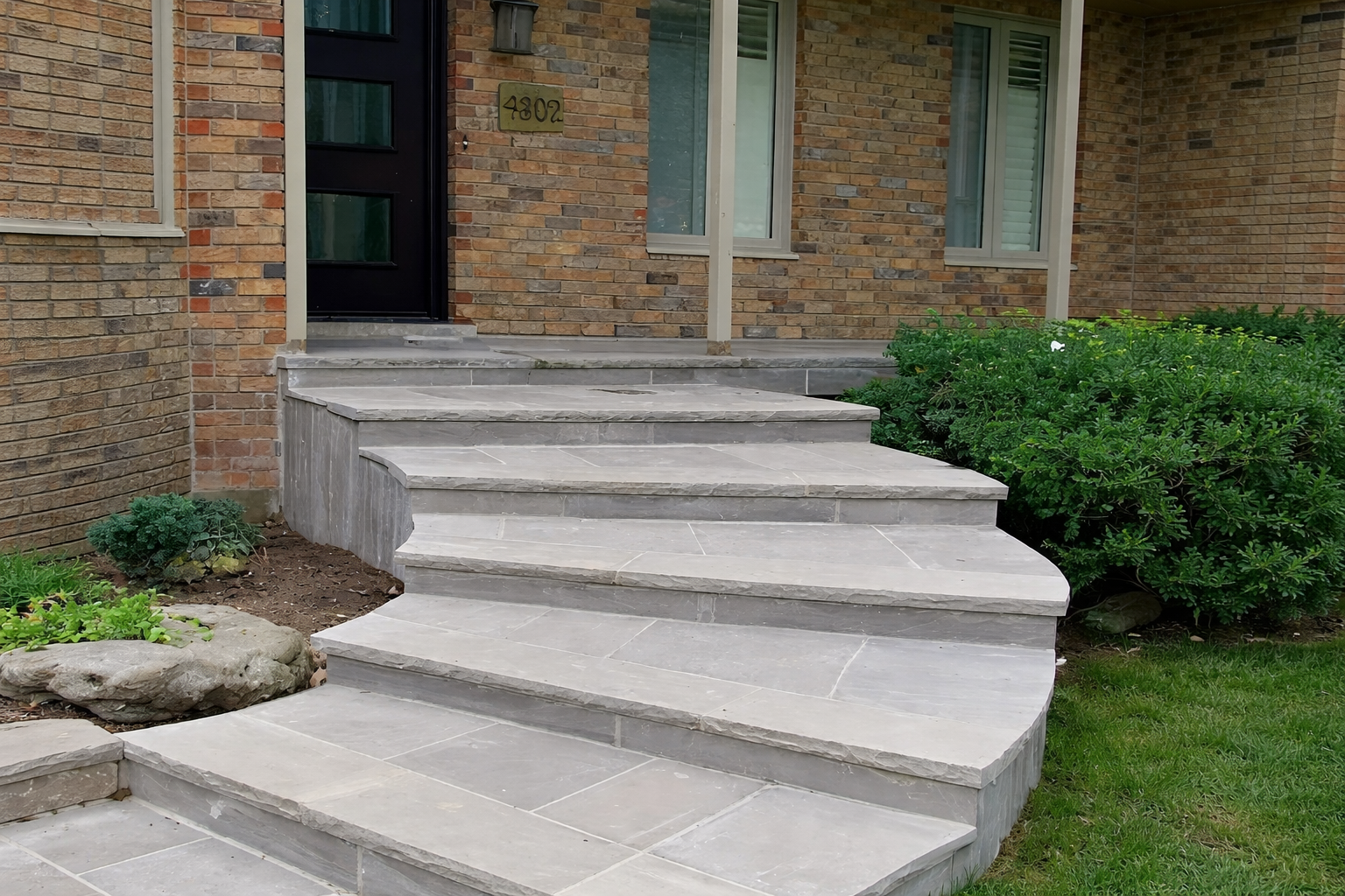 Stone steps leading to a black front door of a brick house, with green plants and bushes on the side.