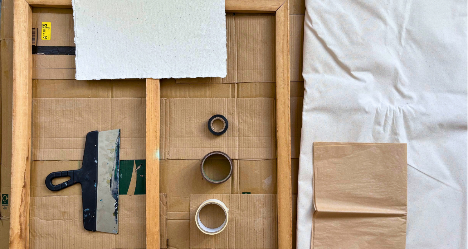 Art materials for surface preparation, including a wooden stretcher frame, cardboard, textured paper, loose canvas, adhesive tapes and a wide paint scraper.